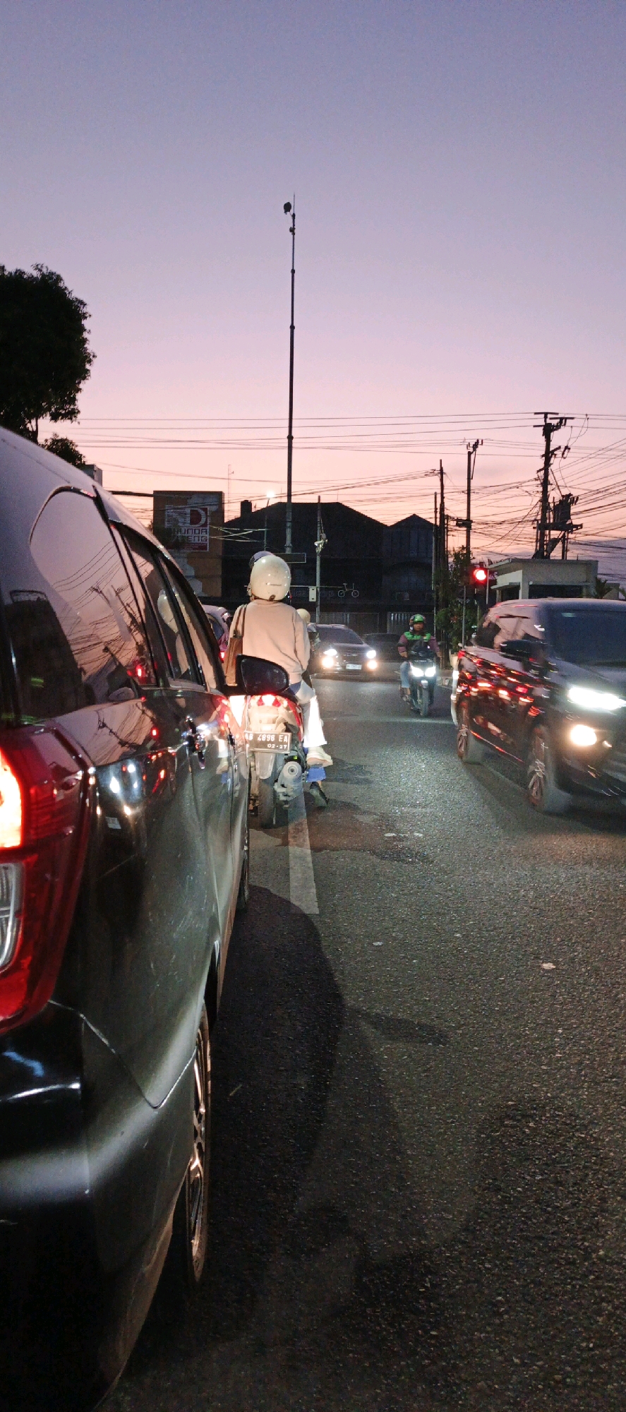 The image depicts a street scene in Yogyakarta, Indonesia, during twilight. The sky is a gradient of purple and pink, indicative of sunset or sunrise. In the foreground, the rear of a black car is visible on the left side of the frame, its taillight illuminated. Beside it, a scooter with two occupants is stopped in traffic. The rider wears a white helmet, and the passenger is seen from behind. The license plate on the scooter reads "AA 4896 EA".
Further down the road, other vehicles are present, including a black car with its headlights on and a motorcycle with a rider in a green jacket. Traffic lights are visible, with one red light illuminated, indicating a halt in traffic. Buildings and power lines are scattered across the background, typical of an urban environment. A tall pole with a camera or sensor is also noticeable. The asphalt road shows signs of wear and tear, with white lane markings. The overall impression is of a busy city street during a period of low light.