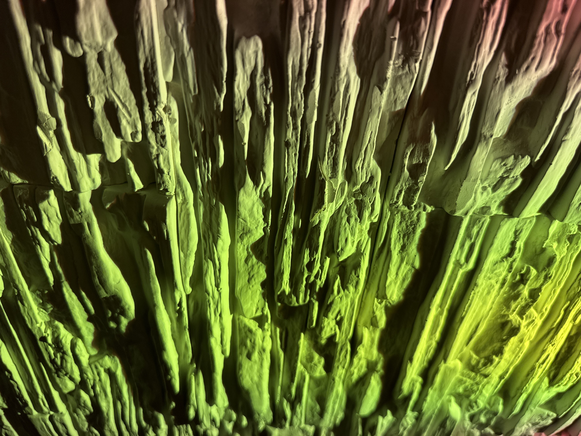 The image presents a close-up, abstract view of a highly textured, vertical surface. The surface is composed of numerous elongated, irregular ridges and grooves, giving it a craggy and somewhat organic appearance, resembling flowstone formations, stalactites, or perhaps a sculpted artistic rendering. The varying depths and forms of these textures create a strong sense of three-dimensionality and depth.
The scene is dramatically illuminated by artificial light sources. The primary coloration is a vibrant lime green and yellowish-green, which covers the majority of the textured surface, particularly pronounced towards the lower and right sections of the frame. Towards the upper left, there are subtle hints of warmer tones, possibly reds or purples, blending into the green. Deep, dark shadows are cast by the prominent ridges and crevices, further accentuating the contours and adding to the dramatic atmosphere.
There are no discernible people, specific objects, or ongoing activities. The focus is entirely on the static, intricate texture of the surface and its illumination. The artificial, colorful lighting suggests an indoor or controlled environment, such as a cave, an art installation, a themed attraction, or a theatrical set, rather than an outdoor natural landscape. No text is visible in the image.