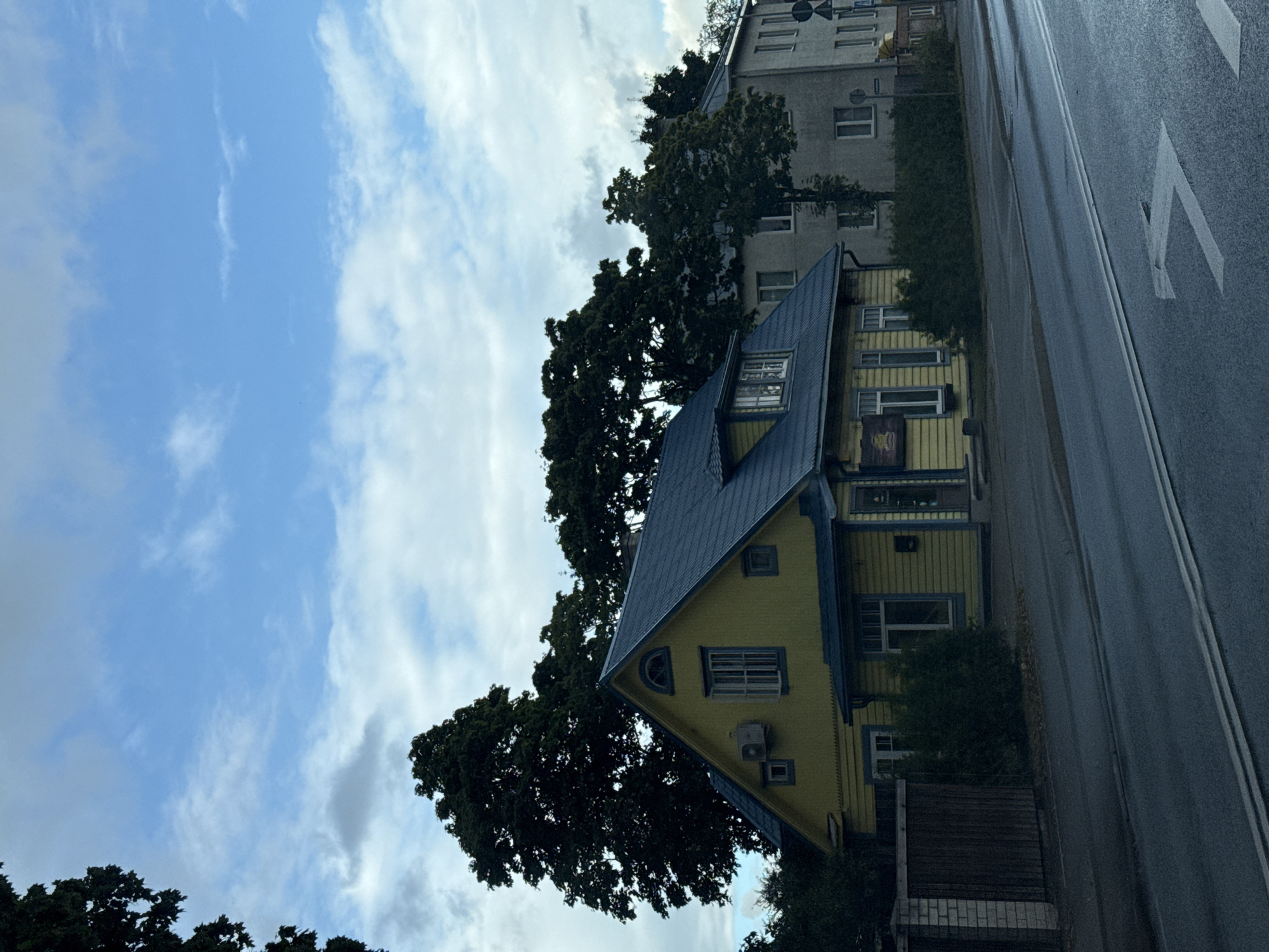 The image shows a yellow, two-story house with a blue roof, located on a street in Tallinn, Estonia. The house has several windows and an air conditioning unit mounted on the exterior wall. To the right of the yellow house is a taller, gray building. Trees are visible in the background, and the sky is cloudy. The street in the foreground is wet, suggesting recent rain. There are white lines painted on the road, indicating traffic lanes. A brick fence and a wooden gate are visible to the left of the yellow house. The overall scene depicts a residential or commercial area in an urban setting.