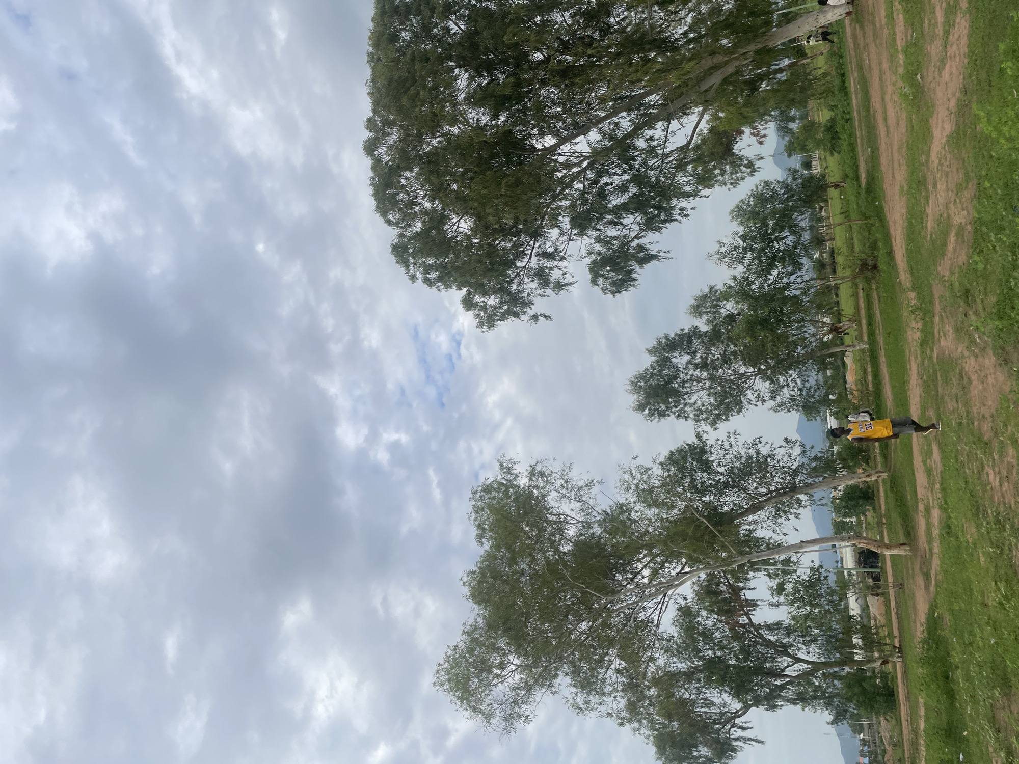 In the city of Jauro Yinu, Nigeria, under a cloudy sky, a young man wearing a yellow sleeveless t-shirt with the number 32 and grey shorts walks across a grassy, dirt-covered area. He carries a white bag over his right shoulder. Several tall trees with sparse green foliage are scattered throughout the scene, some leaning. In the background, there are signs of construction with some buildings visible, and further in the distance, a range of hills can be seen. The ground is uneven with patches of grass and dirt paths. The overall impression is an outdoor setting in a developing area.