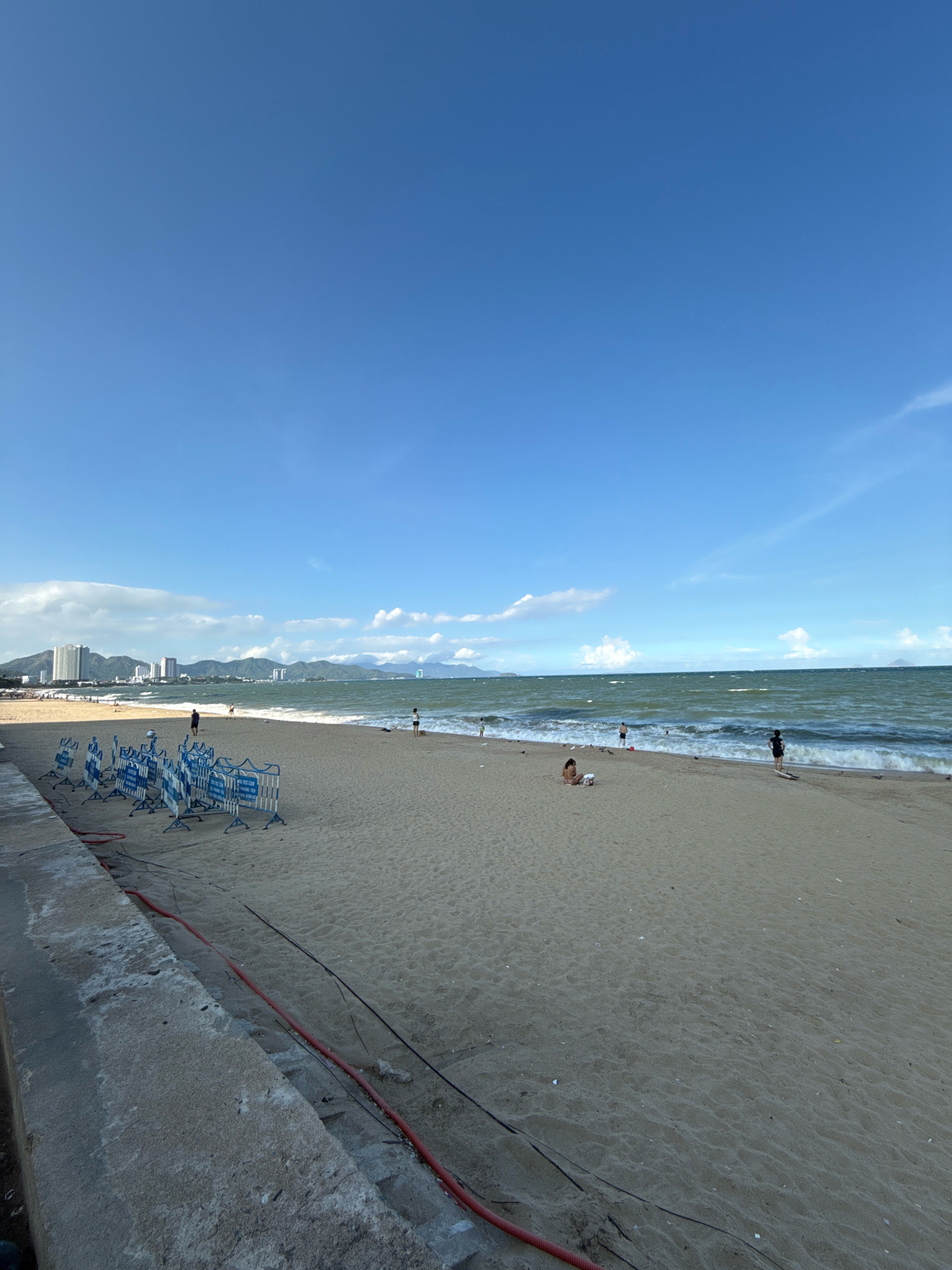 The image depicts a sandy beach in Nha Trang, Vietnam, under a clear blue sky with scattered white clouds. In the foreground, a concrete wall lines the edge of the beach, with red and black cables running along it and onto the sand. Several temporary blue and white barriers, possibly for crowd control or safety, are set up on the sand. Further down the beach, people are visible engaging in various activities. Some individuals are walking or standing near the waterline, while others are seated or lying on the sand. The sea is visible in the background, with moderate waves and a distant shoreline featuring mountains and buildings, characteristic of Nha Trang's coastal cityscape. The overall scene is one of a relatively populated beach on a sunny day.