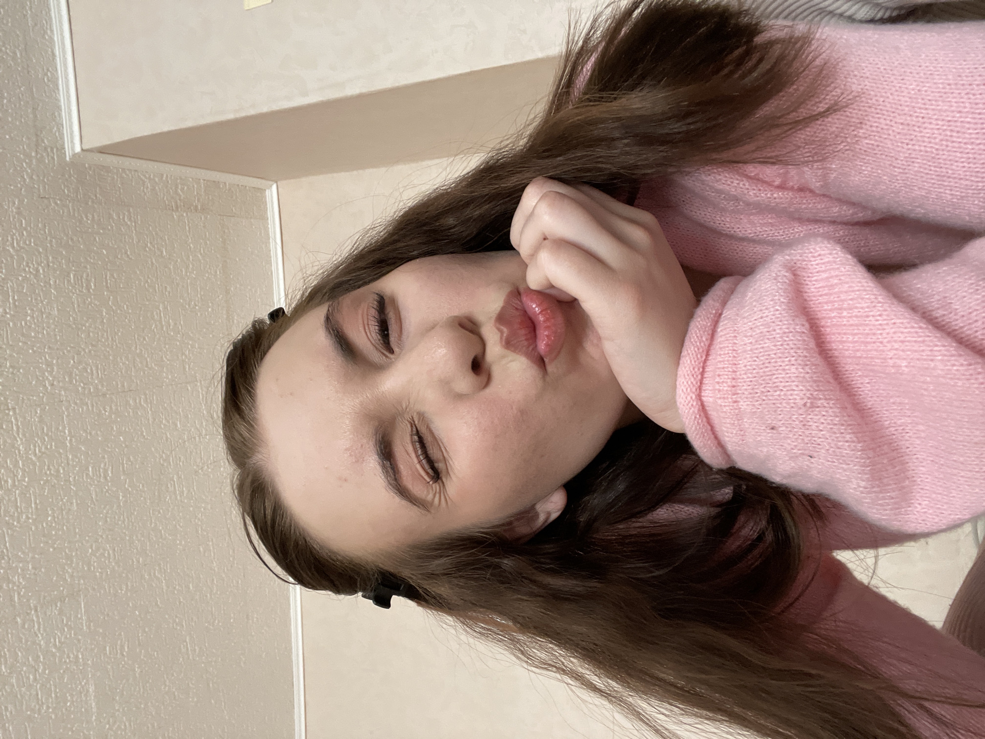 A young woman with brown hair and fair skin, wearing a pink sweater, poses with her eyes closed and lips pursed. She rests her face on her hand. The background is a cream-colored wall. The location is unknown and the questioner's claim is unverifiable.