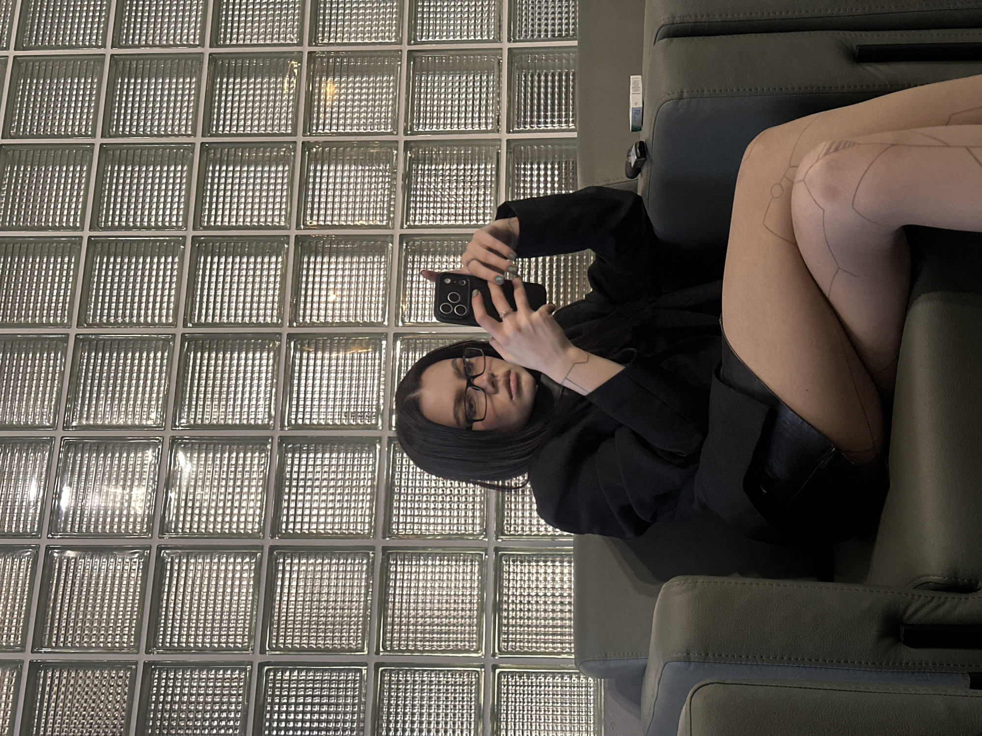 A young woman with dark hair and glasses sits in a green armchair and holds a smartphone to take a photo. She wears a black jacket and patterned tights. Behind her is a wall made of glass blocks. The image was taken indoors. It is impossible to confirm this location is in Moscow, Russia, using only visual data.