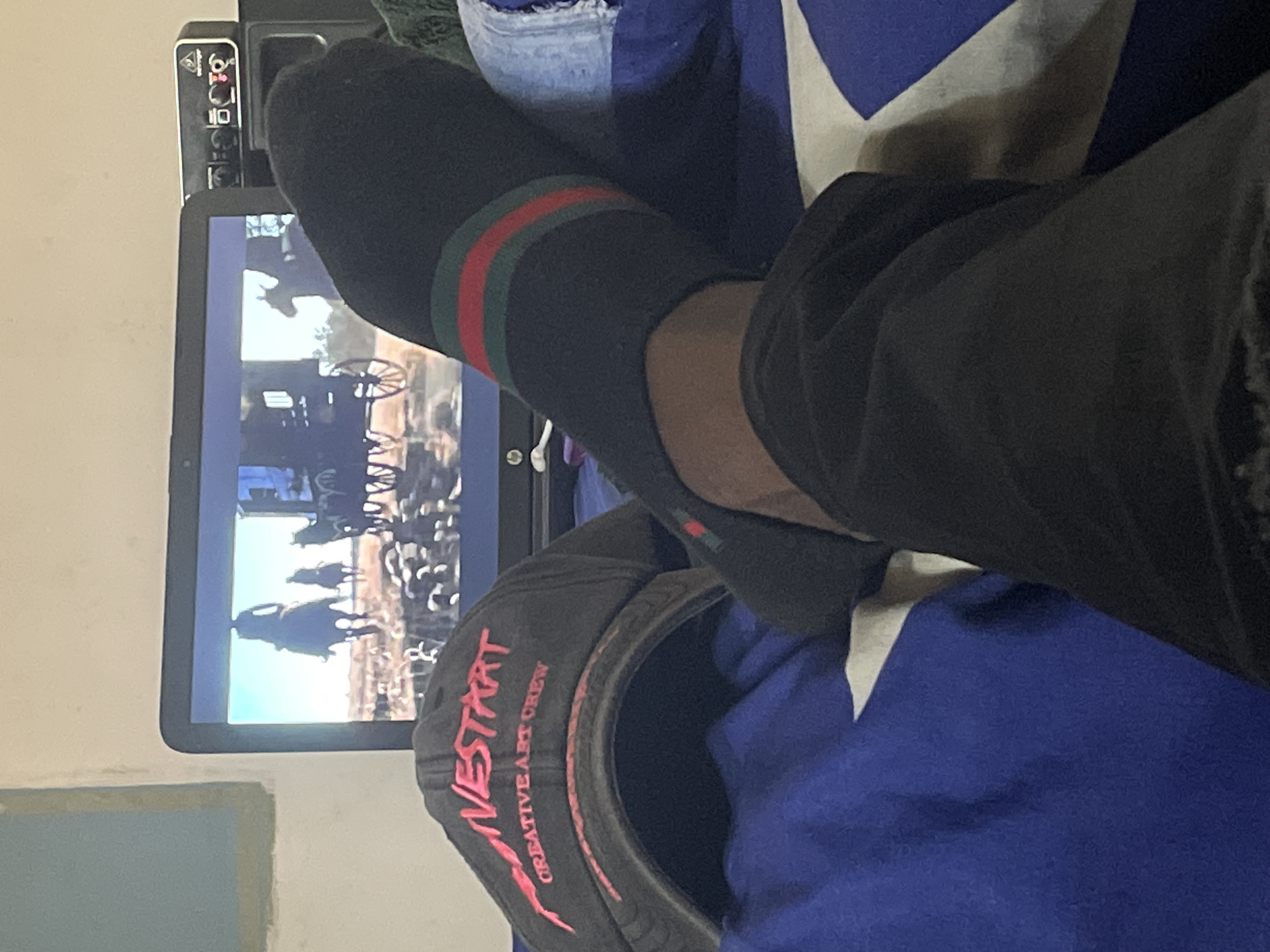 The image shows a person wearing a black sock with green and red stripes, resting their foot near a laptop screen displaying a scene with horses and a carriage. A baseball cap with the words "WESTART CREATIVE ART CREW" in pink is also visible. The person is sitting on a blue and white patterned surface. The location is unknown and there is no evidence the photo was taken in Jos, Nigeria.