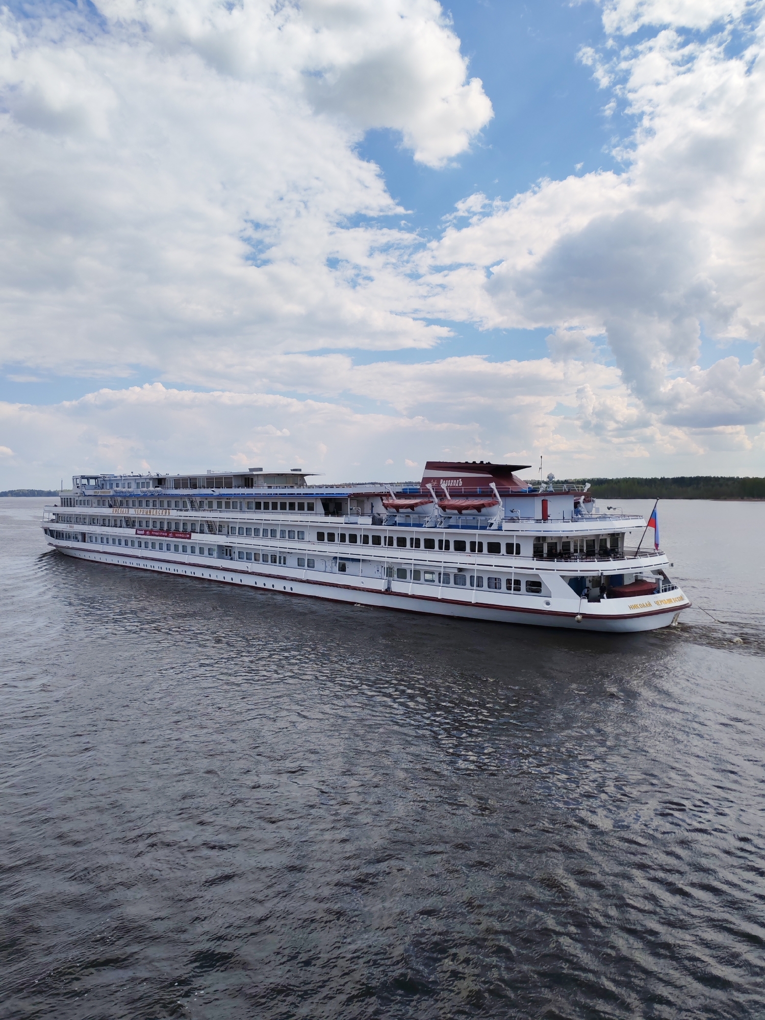 The image shows a white river cruise ship, the "Nikolai Chernyshevsky," sailing on a dark river under a cloudy sky. The ship has multiple decks and a red and white section at the top rear. A Russian flag is visible at the stern. The river reflects the sky and the ship. In the background, the far bank of the river has a line of trees. The exact location in Pruzhinino, Russia, cannot be determined with the provided image. There are no people visible in the image.