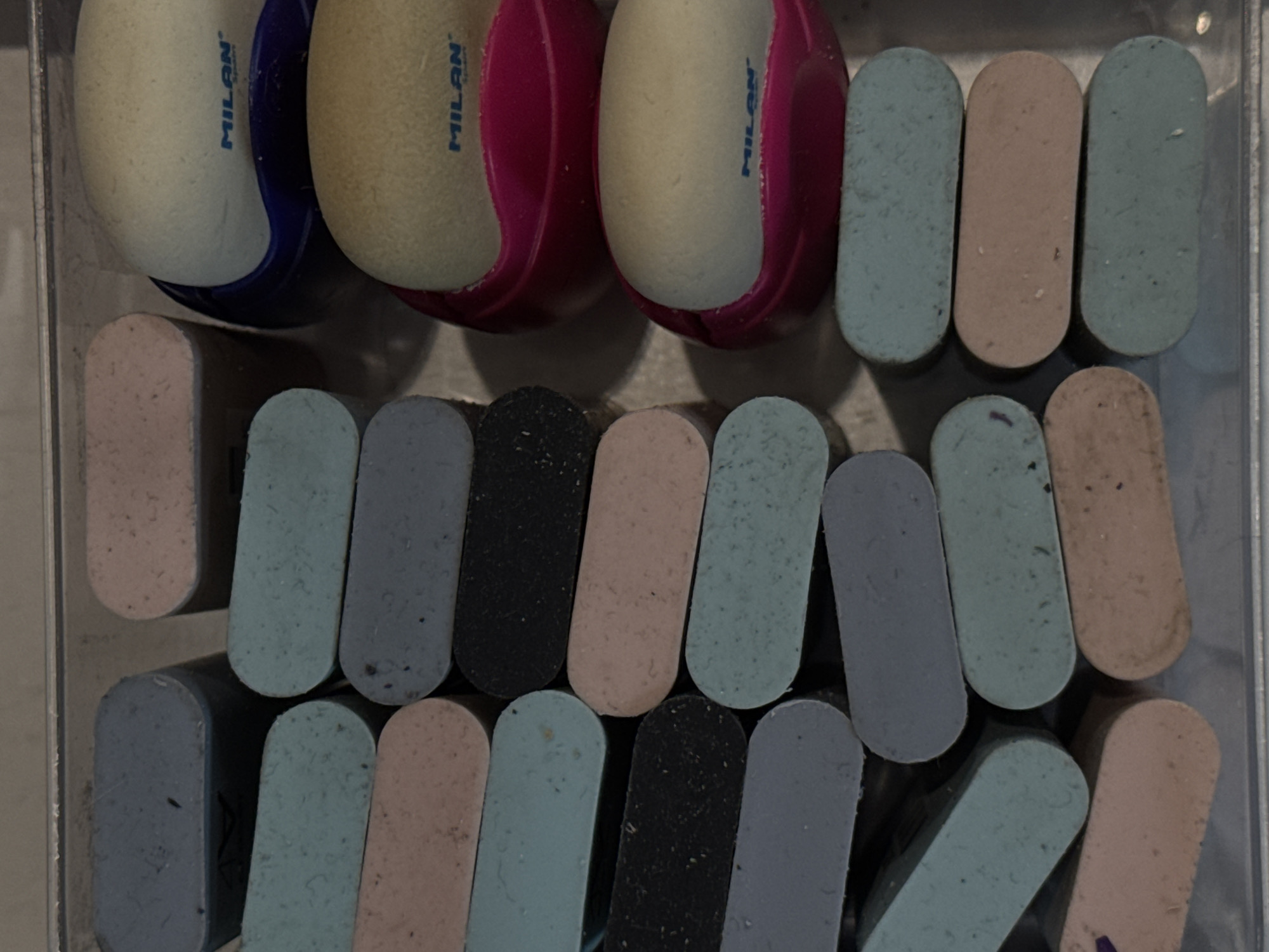 The image shows a close-up, overhead view of a metal container filled with a collection of rectangular erasers and two round erasers housed in pink and blue plastic holders. The erasers are arranged in vertical stacks. The rectangular erasers are in various pastel colors, including light blue, pink, purple, and black. The round erasers appear to be white with blue and pink branding. The container is open at the top and appears to be a shallow tray. The lighting suggests an indoor setting. The image does not contain any visual information that would allow for the identification of a specific location in Moscow, Russia.
