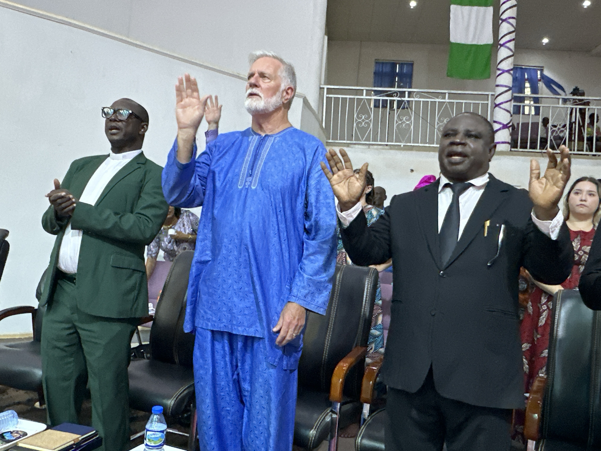 In Yelwa, Nigeria, a man in a green suit and glasses stands on the left with his hands clasped, looking upwards. To his right, a man with a white beard, wearing a blue traditional outfit, also looks upwards with his hands raised. In the center, a man in a black suit and tie faces forward with his right hand raised. A Nigerian flag hangs in the background. Several people are visible in the background, some seated and others standing. The scene appears to be indoors, possibly in a church or community hall.