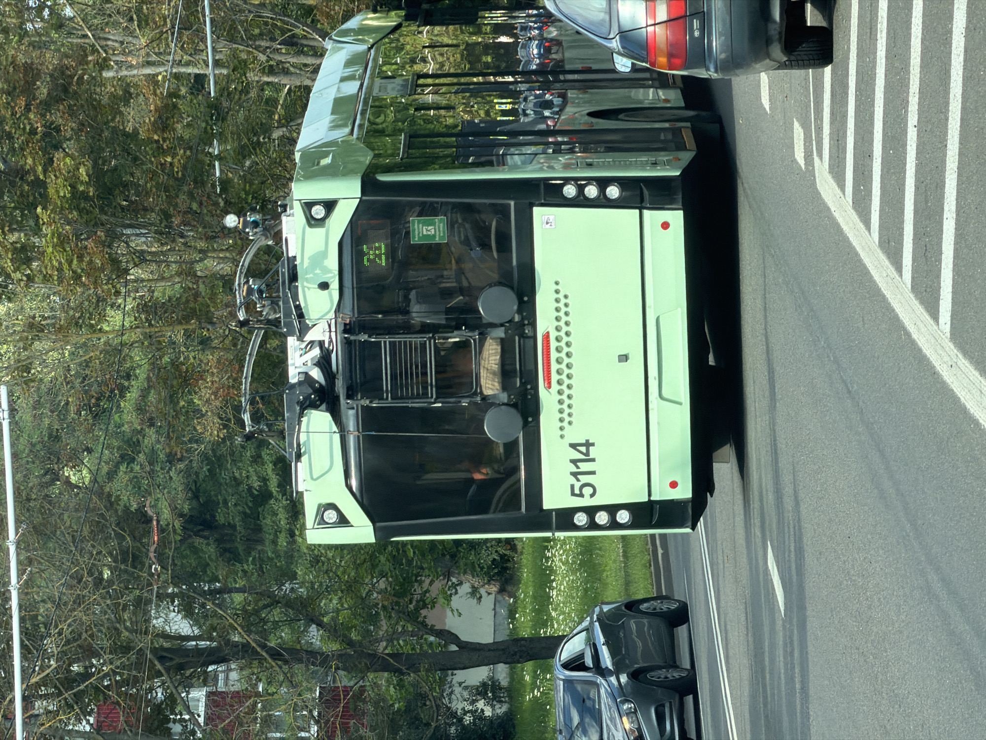 A light green trolleybus with the number 5114 prominently displayed on its side is parked on a paved road. The trolleybus has a modern design with large tinted windows, reflecting the surrounding trees and sky. It has several lights on the front and rear. A dark gray car is parked behind the trolleybus, also on the road. The scene appears to be in an urban or suburban area, with trees and green grass visible on the side of the road. The image is taken during the day, with sunlight casting shadows. There is a green sign with white text on the rear of the trolleybus. The image is rotated 90 degrees counterclockwise.