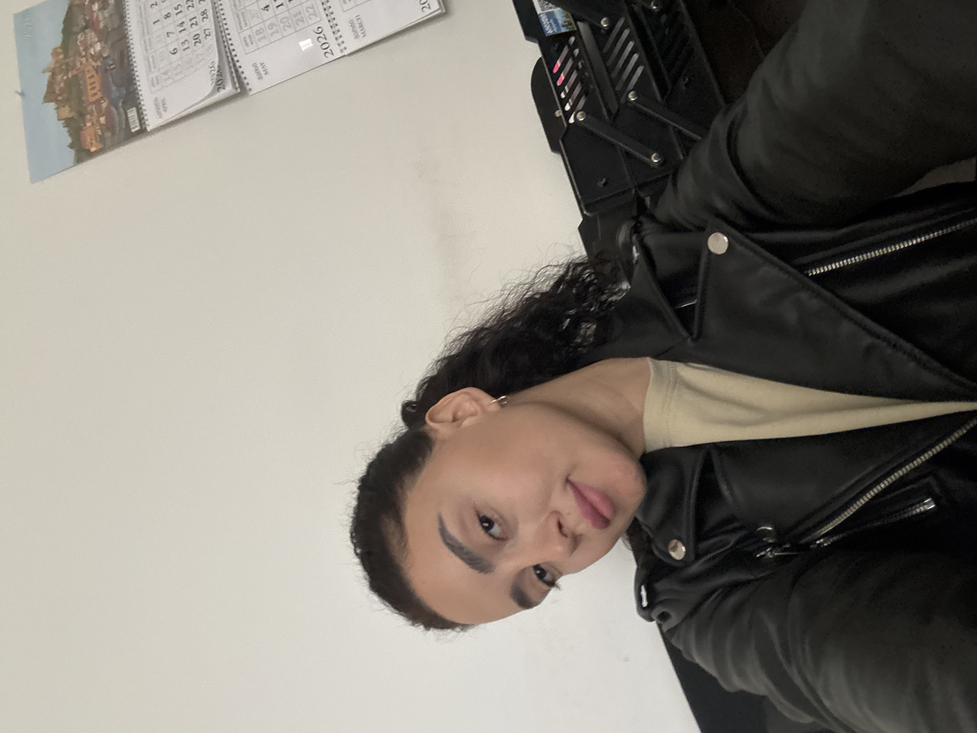 The image shows a woman wearing a black leather jacket. She has dark, curly hair pulled back, fair skin, and visible earrings. She is looking towards the camera with a slightly playful expression, sticking out her tongue.
Behind her, a black metal object is visible, likely part of some equipment or machinery. A calendar hangs on the wall to her left, with the year 2026 visible. The background appears to be a simple, light-colored wall.
Without further information or context, the specific location in Tbilisi, Georgia cannot be determined.