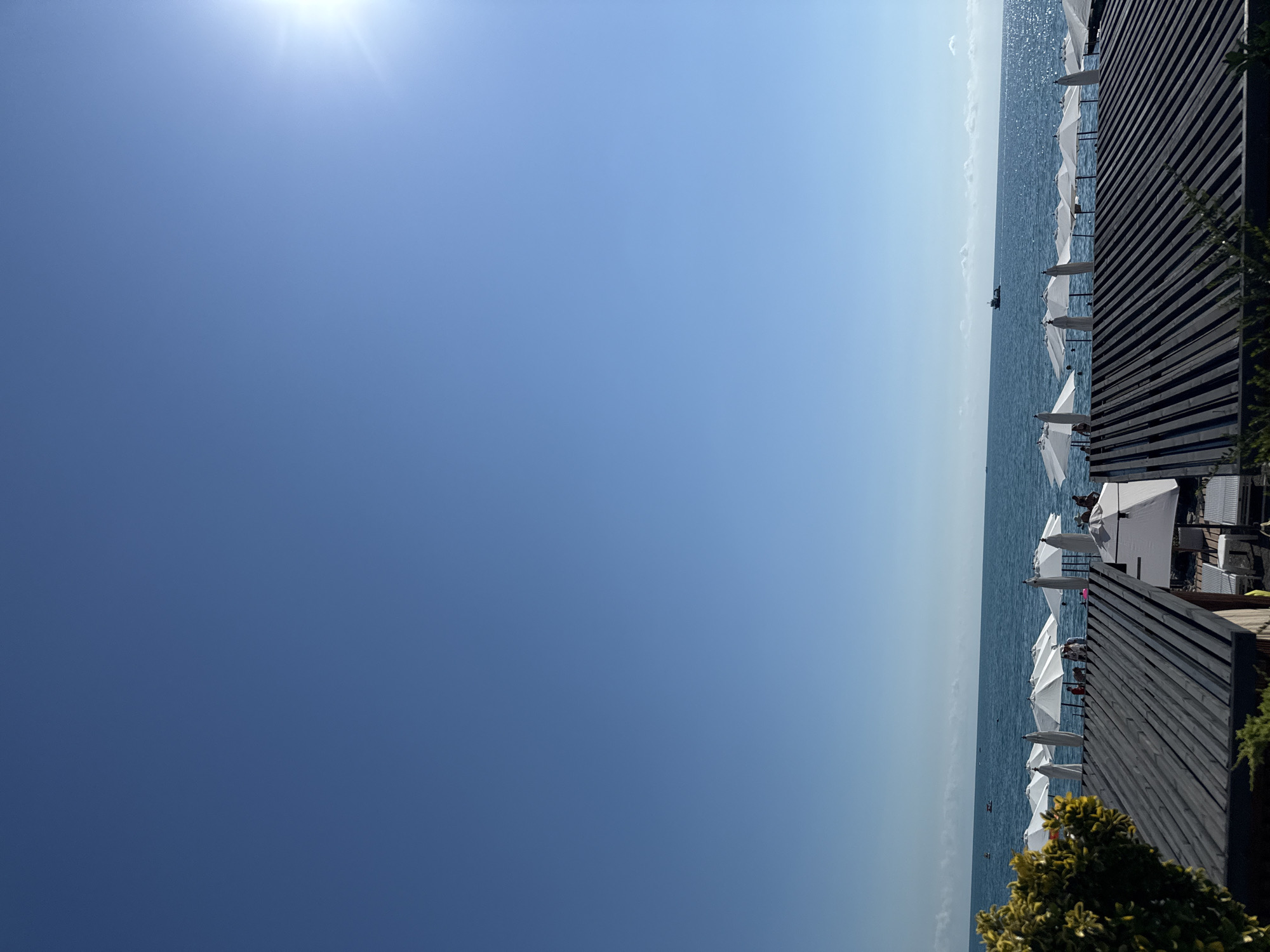 This media file depicts a seaside resort scene, likely in Sochi, Russia. The image is oriented vertically, with the bright sun and a clear, vast blue sky dominating the left two-thirds of the frame. The sun appears high in the sky, suggesting midday. The right side of the image features a wooden structure, possibly a deck or boardwalk, with a railing. Lined up along this structure are numerous white beach umbrellas, many of which are open, providing shade. Beneath some of the umbrellas, people can be seen seated, relaxing. A narrow strip of the blue sea stretches horizontally across the upper right quadrant of the image, its surface shimmering under the sunlight. A small boat is visible in the distance on the water. The overall mood is one of a bright, sunny day at a beach resort, with a sense of leisure and relaxation. There is no visible text in the image.