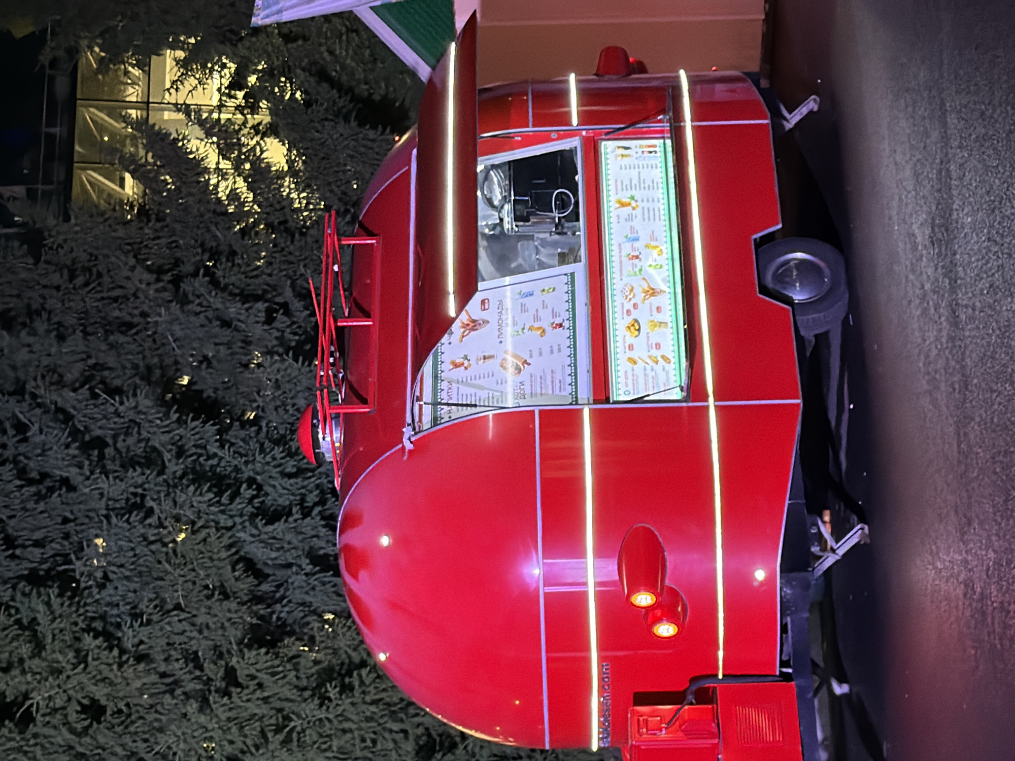 In an outdoor setting at night, a bright red, retro-style food truck is parked on an asphalt surface. The food truck has a rounded, domed roof and a serving window that is partially open, revealing a menu board. The menu board displays images of food and drinks, including what appear to be hot dogs, fries, and various beverages. The truck is decorated with horizontal white lights along its exterior. In the background, dense, dark green evergreen trees are visible, with hints of yellow light filtering through their branches, suggesting a building or artificial lighting behind them. The overall scene appears festive, possibly at an event or market.