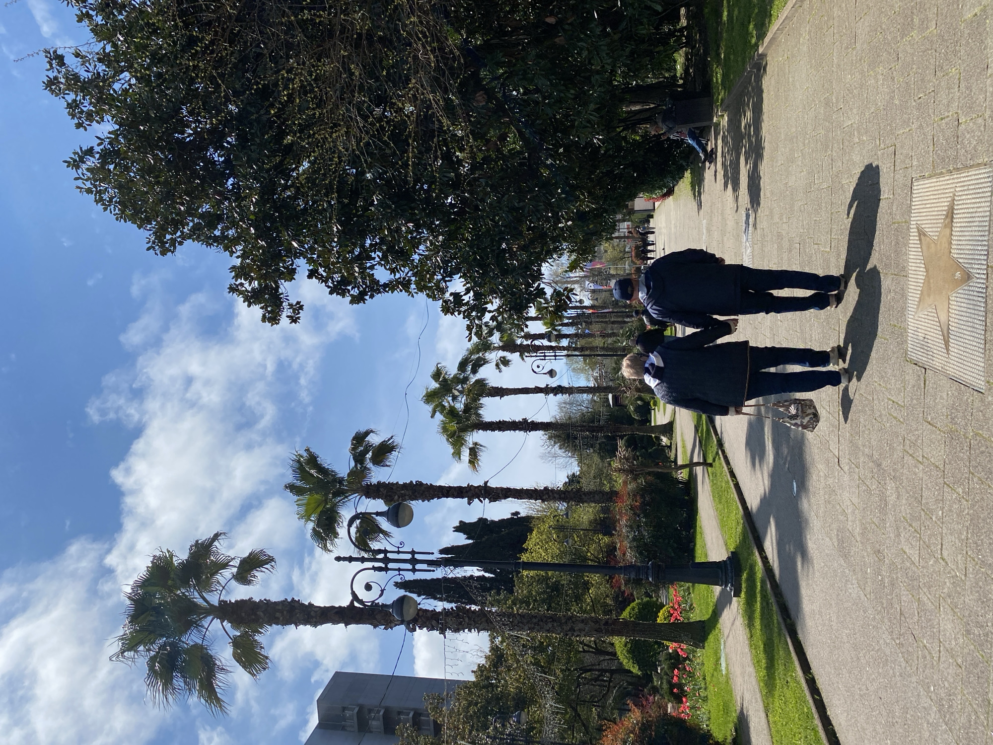 Two people walk along a paved walkway. A large tree and tall palm trees line the walkway, along with lamp posts and flower beds. A building is visible behind the palm trees. A star-shaped plaque is embedded in the walkway. The sky is blue with scattered clouds. This location is likely the Alley of Stars in Sochi, Russia.