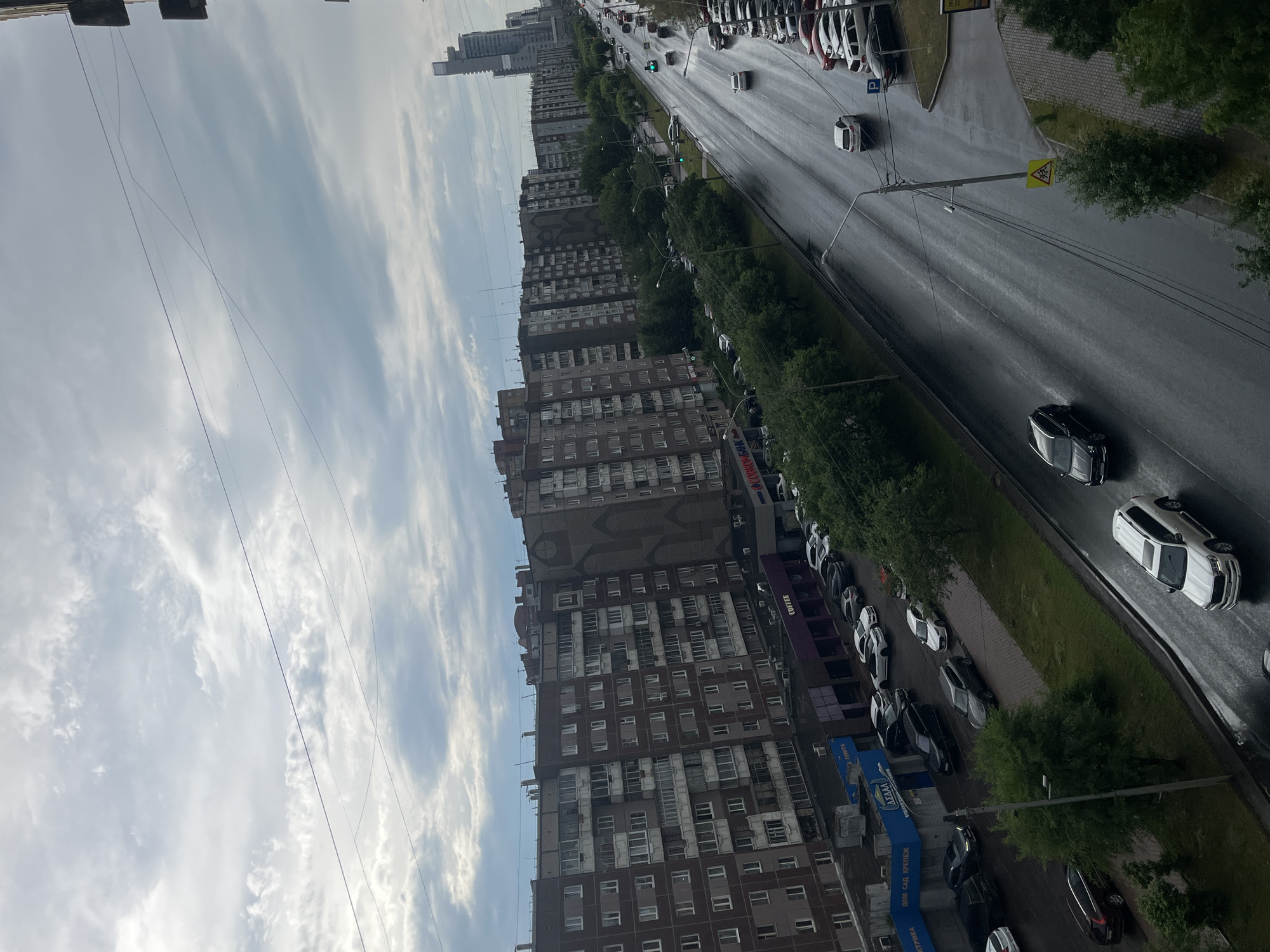 The image depicts a street scene in Krasnoyarsk, Russia, under a cloudy sky. The foreground shows a wet asphalt road with several cars driving and parked. On the left side of the road, there are multi-story apartment buildings with balconies and windows. Some of these buildings have storefronts on the ground level, including a sign that reads "COFFEE" and another with Russian text. Trees line the sidewalk and median of the street. In the distance, more apartment buildings and a tall, modern skyscraper are visible. Traffic lights are present, with some showing green. The overall atmosphere is overcast, suggesting potentially rainy or post-rain conditions, as indicated by the wet road surface.