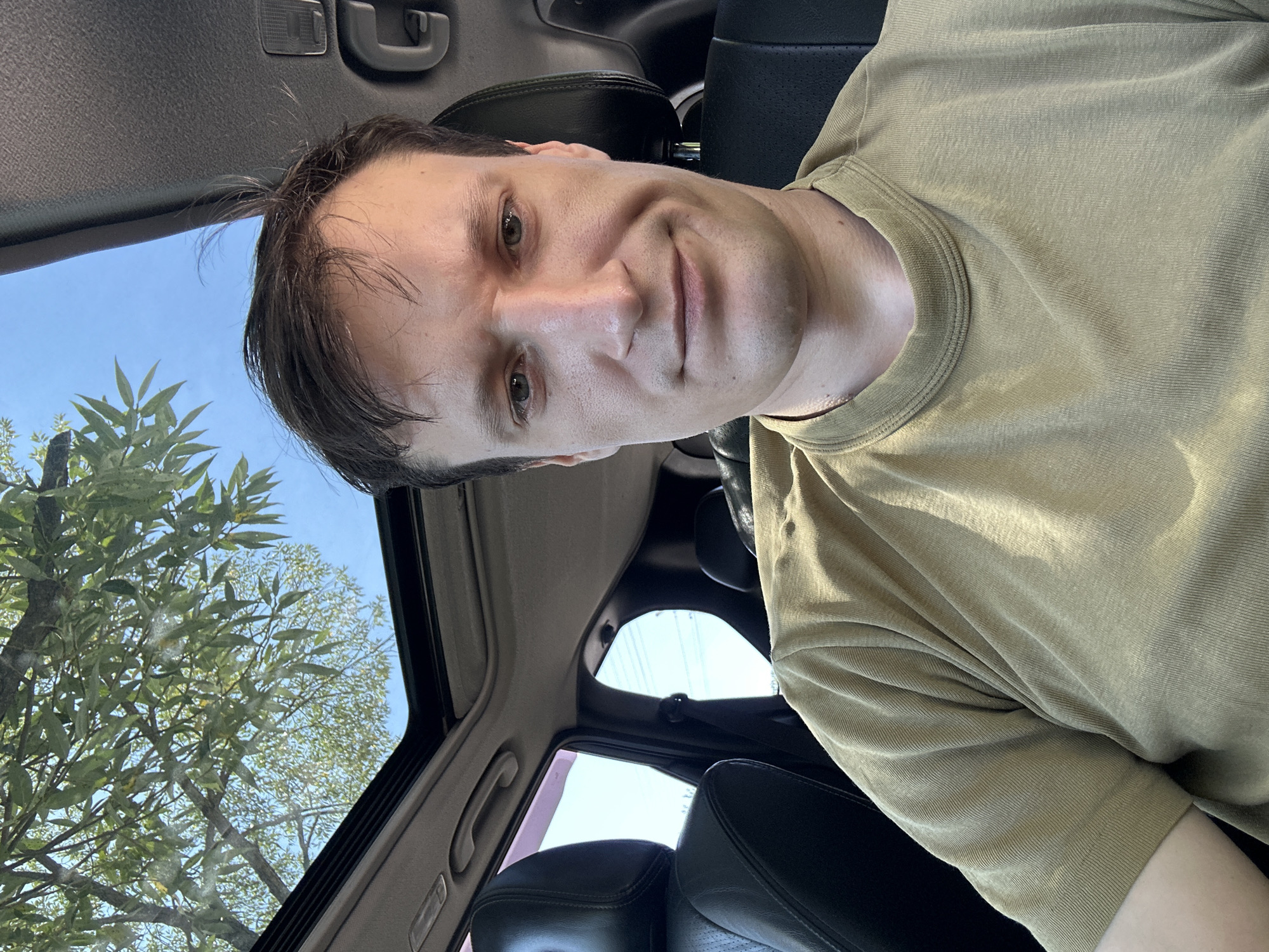 The image shows a young adult male in the driver's seat of a vehicle, looking at the camera. The man has short, dark brown hair and light-colored eyes. He is wearing a light brown t-shirt. The car has a sunroof, through which trees and a bright blue sky are visible. The interior of the car appears to be black leather. The location is identified as Makiv, Ukraine.