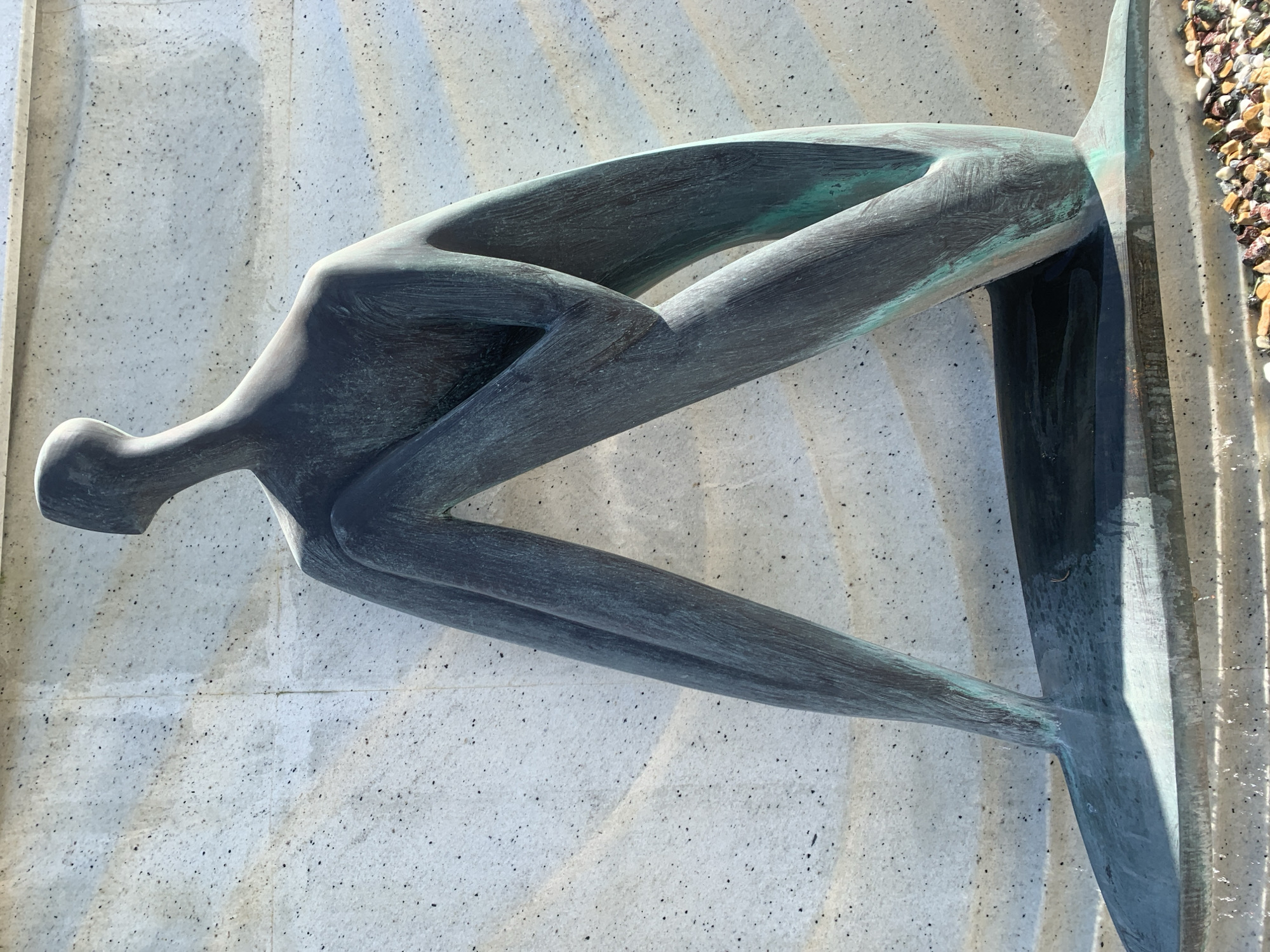 This is a medium shot, taken from a slightly low angle, of a bronze sculpture of a seated, stylized human figure against a textured concrete wall. The sculpture is dark green with hints of blue-green patina and appears to be made of a smooth, polished metal. The figure is abstract, with elongated limbs and a simplified head. It is seated with its legs drawn up, forming a triangular shape. The base of the sculpture is also made of the same bronze-like material and rests on a textured surface, possibly concrete or stone, with a scattering of small, colorful pebbles at the bottom of the frame. The background wall is light gray and features a repeating wavy pattern, with visible speckles of dark material embedded in the concrete. The lighting suggests it is daytime, with shadows cast by the sculpture on the base and the wall. The sculpture is a public artwork, likely located in an outdoor space in the city of Lviv, Ukraine.
