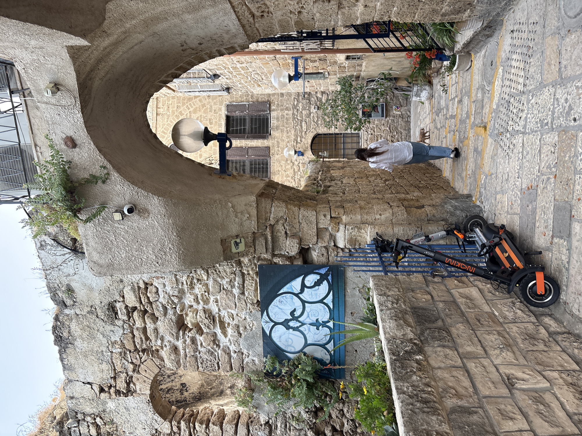 The image depicts a narrow alley in Old Jaffa, Tel-Aviv, Israel, characterized by stone architecture. The scene includes a stone archway and building facades. A woman in a white shirt and jeans is walking away from the camera. An orange and black electric scooter is parked on the side. A blue painted canvas artwork of an abstract floral design is attached to a wall near the ground, and the stone buildings also feature mounted light fixtures.