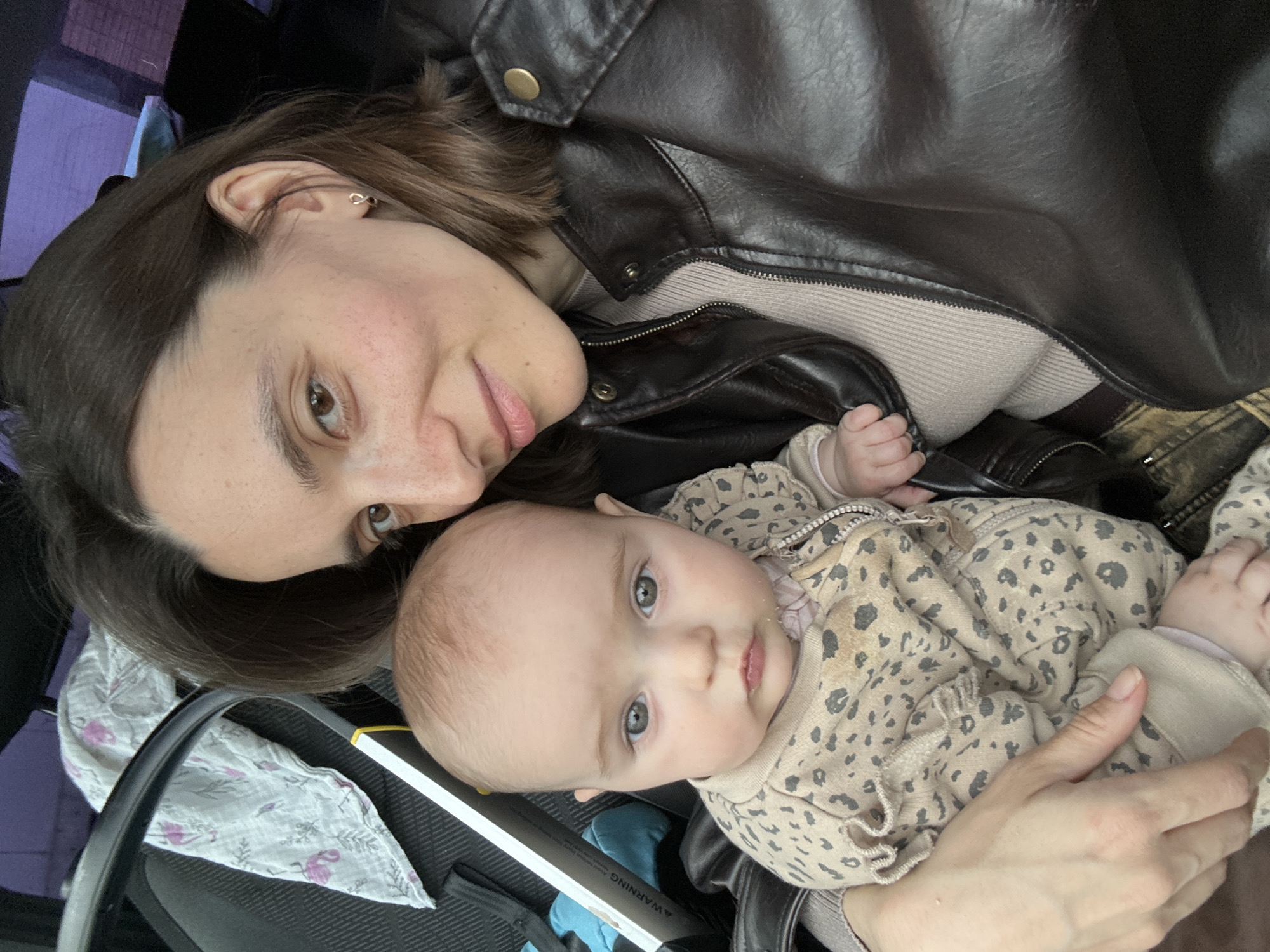 A woman with dark brown hair and eyes, wearing a brown leather jacket, is holding a baby in her arms. The baby, dressed in a light-colored outfit with a small animal print, is looking towards the camera. The woman is also looking toward the camera. The scene is framed as a close-up shot, with details suggesting a setting in a stroller, possibly in Moscow, Russia, though specific landmarks are not visible.