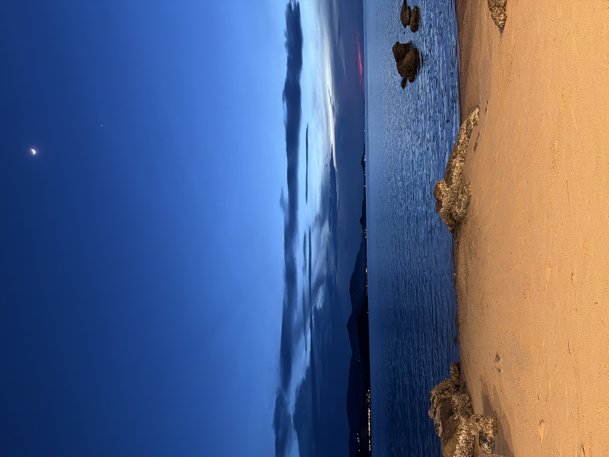 The image depicts a serene twilight scene at a beach in Baan Bo Phut, Thailand. The sky is a deep, rich blue, transitioning into lighter hues near the horizon, where streaks of clouds are illuminated by the fading sun. A crescent moon is visible in the upper left portion of the sky, accompanied by two small, bright dots, possibly stars or planets.
The foreground is dominated by a sandy beach, with rough, textured rocks jutting out at the waterline, some of which appear encrusted with barnacles or similar marine growth. The ocean stretches out to the right, its surface rippling gently, reflecting the twilight sky. In the distance, along the horizon, a faint line of lights suggests a coastal town or settlement.
The overall mood of the image is peaceful and tranquil, evoking the calm of dusk. The colors are subdued, with the deep blues of the sky and sea contrasting with the warm tones of the sand. There is no visible text in the image. The scene suggests a quiet moment of natural beauty, possibly after sunset.