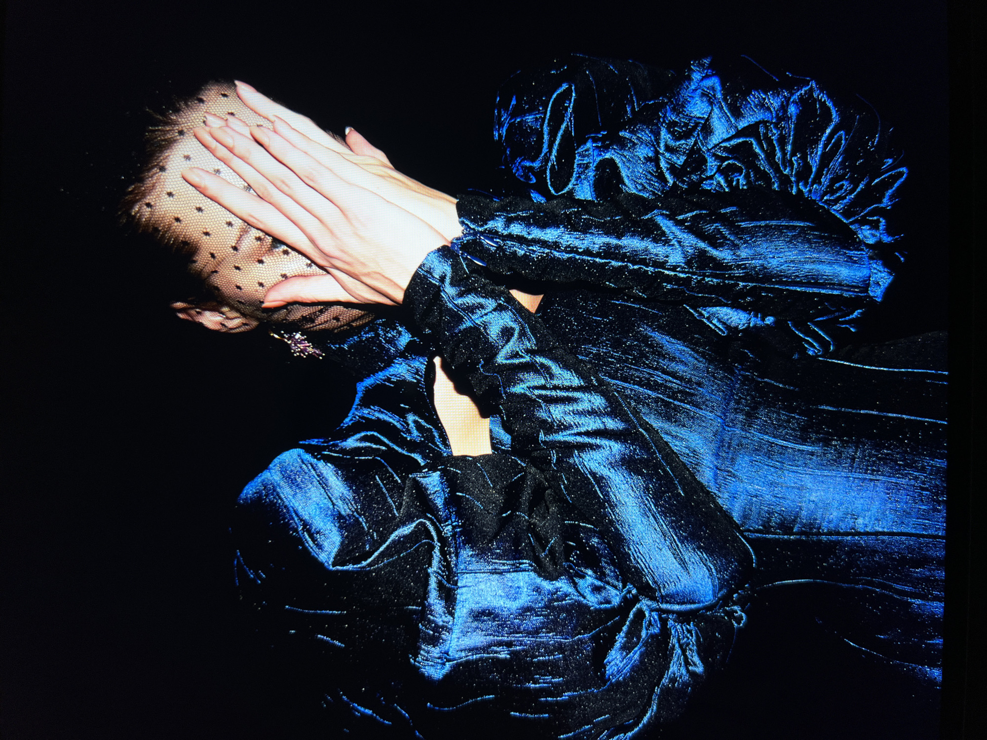 A person, their face obscured by a black dotted veil and hands, is depicted against a dark background. The individual is wearing a voluminous, shimmering blue garment with textured fabric that appears to be a dress or jacket. The garment has prominent ruffles or pleats, particularly around the shoulders and sleeves, creating a dramatic silhouette. The lighting highlights the metallic sheen of the fabric, with strong blue and black tones dominating the scene. The person's hands, with manicured nails, are clasped in front of their face, and one hand is visible through the veil. A small, glittering accessory, possibly an earring or brooch, is visible on the left side of the head. The overall impression is one of mystery and dramatic presentation.