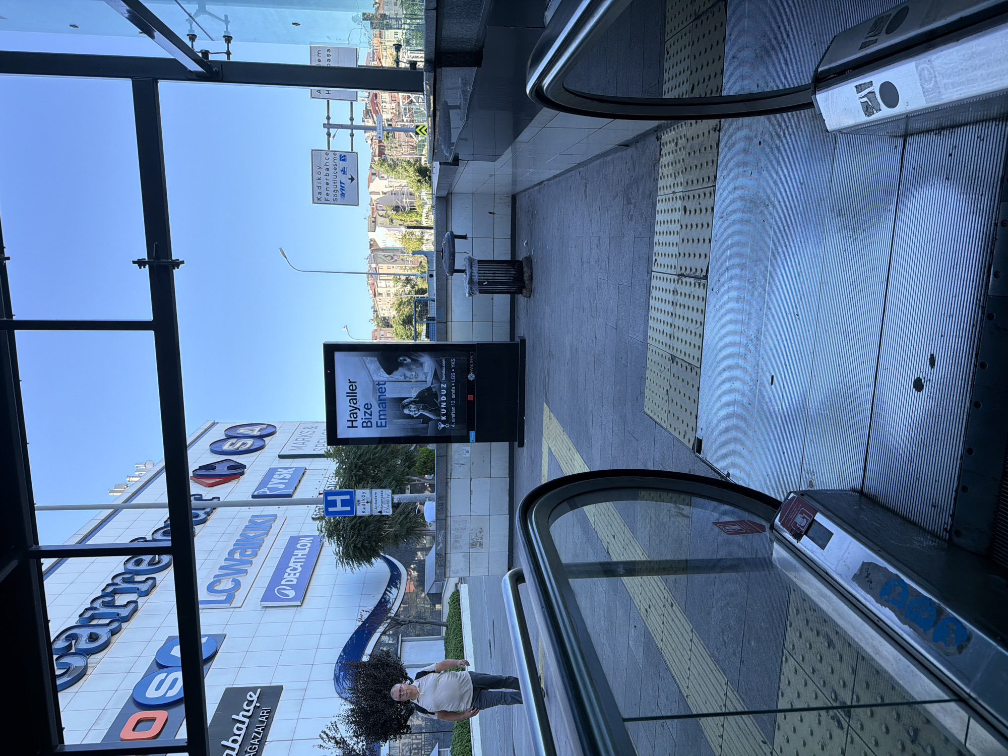 The image shows the entrance to a shopping mall or public transport station in Kadıköy, Turkey. In the foreground, two escalators are partially visible, with metal handrails and textured yellow safety strips on the ground. A man wearing a light-colored polo shirt and dark trousers walks away from the camera on the left side of the frame. Behind him, the facade of a large building is visible, displaying the logos of various businesses including "Carrefour," "LC Waikiki," and "Decathlon." To the right of the man, a tall, illuminated digital billboard displays an advertisement for "Hayaller Bize Emanet" with images of people. Further into the background, a trash can is positioned next to a tiled wall. Above the billboard, traffic signs indicate directions to "Kadıköy," "Fenerbahçe," and "Söğütlüçeşme." The sky is clear and blue, suggesting a sunny day. The overall scene depicts an urban environment with commercial activity and public infrastructure.