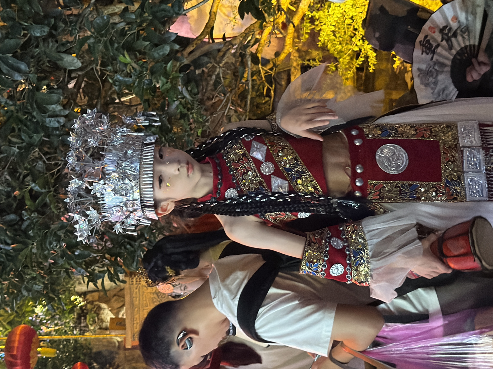 A young girl wearing a traditional elaborate costume and headdress stands in front of lush greenery, illuminated by warm, yellow lighting. She has intricate makeup with glitter on her face. The costume is red and gold, with ornate embellishments and flowing white sleeves. In her right hand, she holds a small red cylindrical object. To her left, a person in a white t-shirt with a black strap is visible, carrying a pink plastic bag. Another person in a gold headdress and dark hair is partially visible behind them. In the background, red lanterns are suspended, and a traditional fan with Chinese characters is held by someone out of frame to the right. The overall scene suggests a cultural event or performance, likely taking place in Yuzhong District, China, given the context of the location.