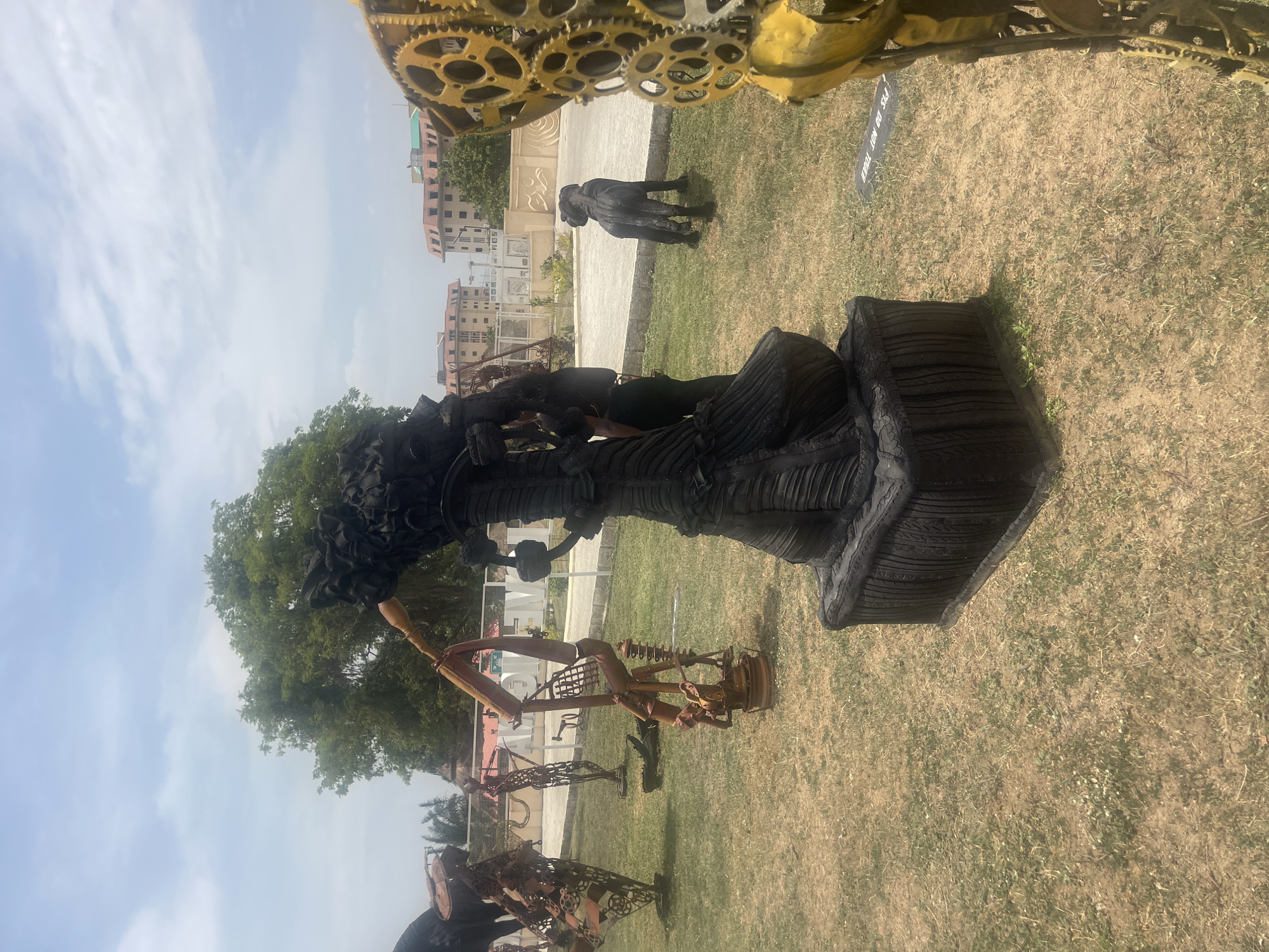 This media file captures an outdoor sculpture park or exhibition under a bright, partly cloudy sky in Abuja, Nigeria. The scene features several large, dark-colored sculptures positioned on a vast grassy lawn.
In the mid-ground, a prominent sculpture depicts a stylized figure with a textured, layered body that widens towards its base, and an elaborate, voluminous headpiece. This piece appears to be crafted from dark, possibly recycled materials such as cut tires or similar industrial components, lending it a distinctive, rugged appearance. A person is visible standing near this sculpture, observing it.
Further into the background, another dark, quadruped-like sculpture, resembling a dog, stands on the grass, with another individual nearby. Additional metallic, abstract sculptures, seemingly constructed from repurposed industrial parts, are scattered across the lawn. The far background reveals multi-story buildings with light-colored facades and reddish roofs, providing an urban context to the open green space. A large tree with dense green foliage stands to the left of the central sculpture.
The upper portion of the image shows a bright blue sky with scattered wispy white clouds, indicating clear weather during the daytime, likely mid-morning or afternoon. A metallic structure with gear-like components, possibly part of another large sculpture or a framing element, is visible in the top right corner. On the ground, near the base of one of the sculptures, a black sign with white text clearly reads: "PLS DO NOT TOUCH." The overall setting suggests a public space dedicated to art appreciation.