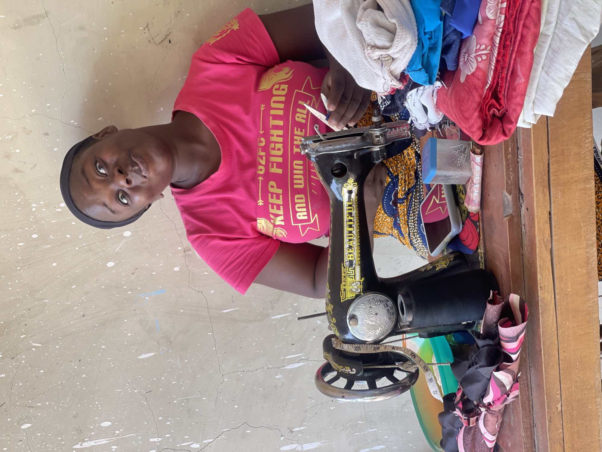 A woman with dark hair and a black headband, wearing a pink t-shirt that reads "GZFC KEEP FIGHTING AND WIN THE ALL", is positioned behind an old, black sewing machine on a wooden table. The sewing machine has the word "BEAUTIFUL" printed vertically on its side. A measuring tape is coiled around the machine's wheel. Various fabrics and materials, including folded cloths in red, white, and blue, are piled on the table near the sewing machine. A small container of pins and a cell phone sit near the machine. The woman holds a small pair of scissors near the sewing machine's needle. Scraps of fabric are bunched on the table's edge. The location is Akwanga, Nigeria, implied by the style of the clothing. The wall behind the woman is a light beige color with scattered white paint spots and visible cracks.