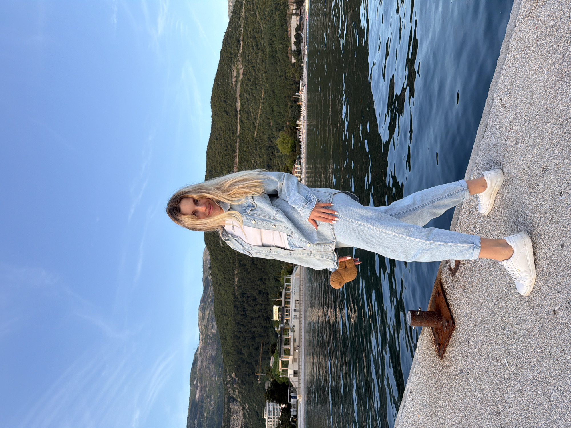 A young woman with long blond hair, wearing a matching light-blue denim jacket and jeans, and white sneakers, is sitting on a concrete pier. She has a small, brown teddy bear in her hand. In the background is the blue water and a green, mountainous landscape. The sky is clear and blue. The location is in the city of Budva, Montenegro.