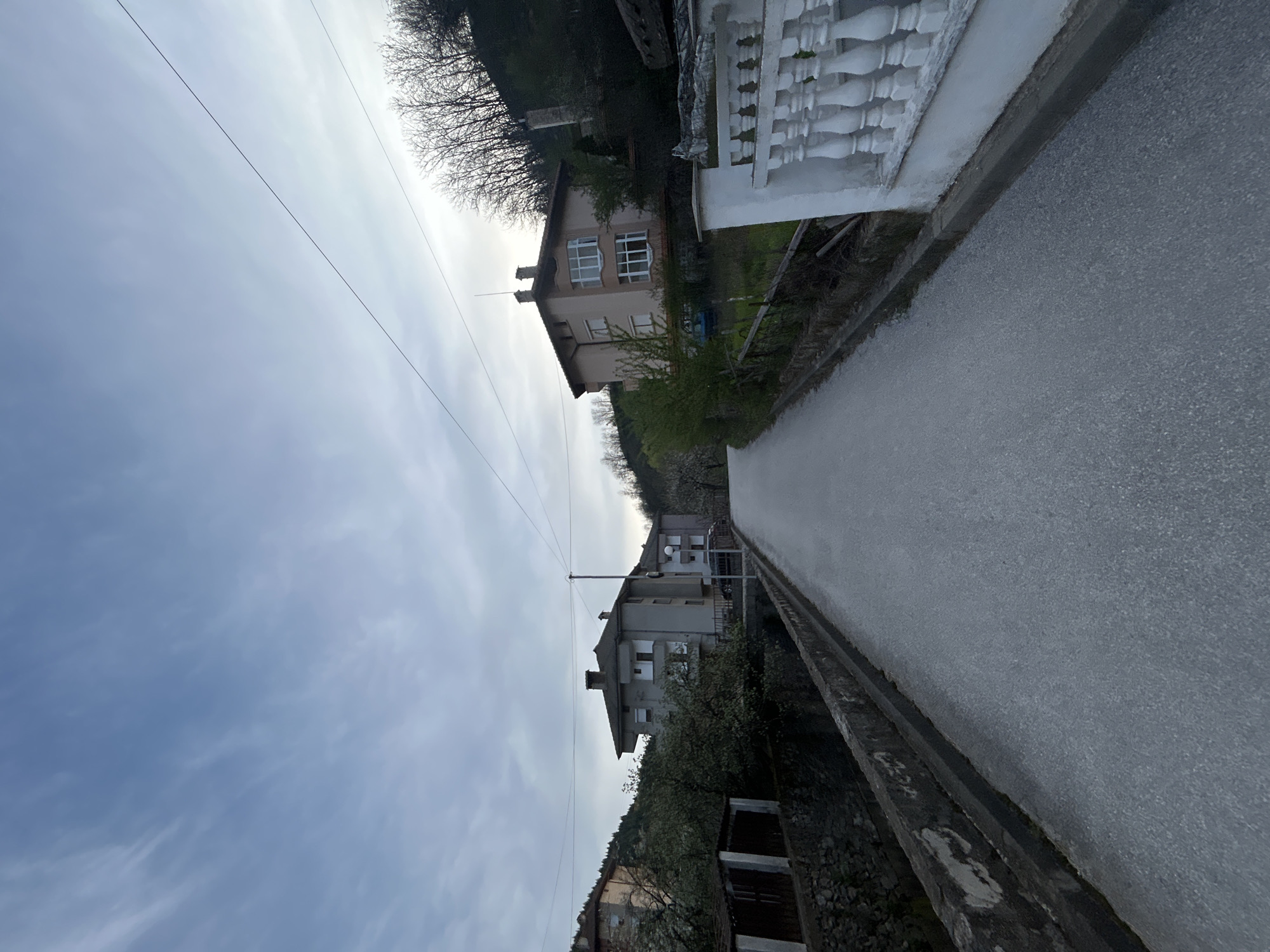 The image shows a narrow asphalt road in Zlatograd, Bulgaria, flanked by houses and retaining walls. On the right side of the road is a white balustrade wall. Above this wall is a residential building with light-colored exterior and white-framed windows. Trees and vegetation grow along the edge of the road and on the hillside behind the house. On the left side of the road, there's a retaining wall and a building with a dark roof and light-colored exterior. Power lines run across the sky above the road. The sky is overcast.