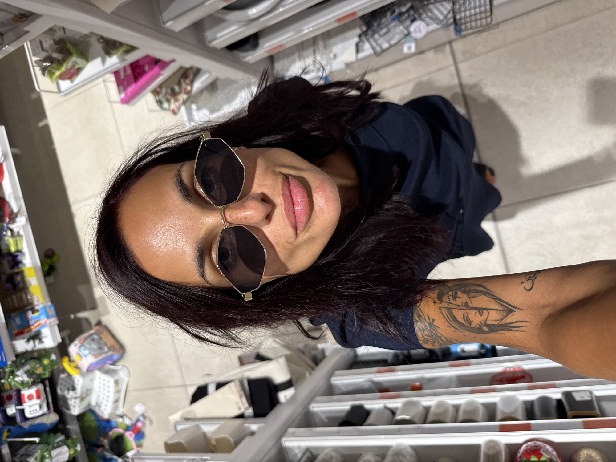 The image is a high-angle selfie taken in a retail store, likely in Pleven, Bulgaria, featuring a woman with dark hair and sunglasses. She is wearing a dark blue dress and has a tattoo on her left forearm depicting two faces split down the middle, with Arabic script below it. She is positioned in front of shelves stocked with various items, including what appear to be household goods or accessories. The store environment is brightly lit with tiled flooring.