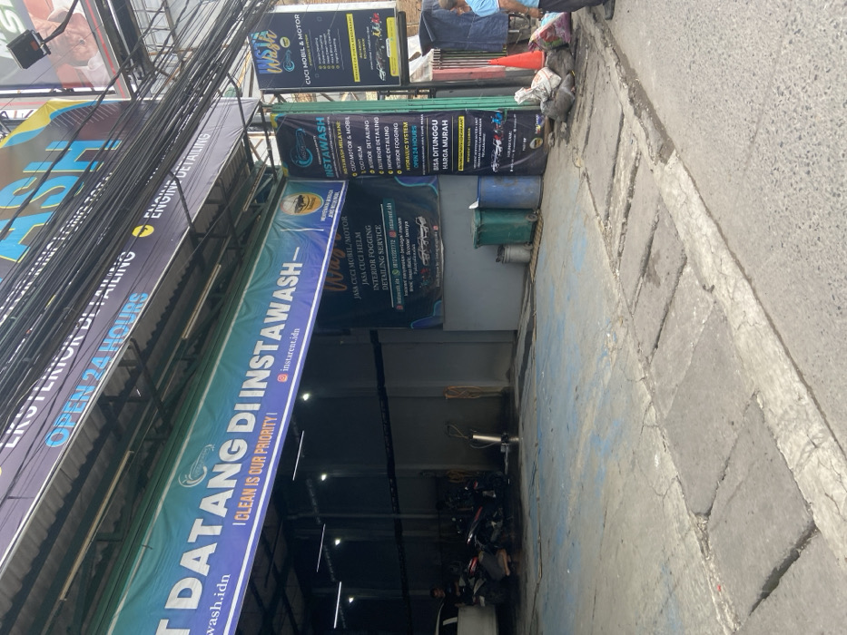 The image shows the exterior of a car wash in Bojongsoang, Indonesia, located at coordinates -6.972, 107.636. The car wash is called "Instawash" and features a large sign with the slogan "Datang di Instawash - I Clean is Our Priority!". The sign is in Indonesian and English. The car wash appears to be open 24 hours a day. There is a smaller sign next to the main one advertising various services including car and motorcycle washing, detailing, fogging, and cleaning. The building has a large open doorway that leads to the car wash area. There is a person in the doorway, but their face is not visible. The person appears to be wearing a light blue shirt. There are several motorcycles parked in the car wash area. A green trash can with a black trash bag is visible in the image. There is a person standing to the right of the trash can, wearing a blue shirt. There is a small orange traffic cone nearby. The ground in front of the car wash is paved with concrete. There are signs with a yellow background and black letters. The signs mention various services offered by the car wash. The image is taken during the day, and there is no sign of rain. There is a building behind the car wash with a "ASH" sign on it.