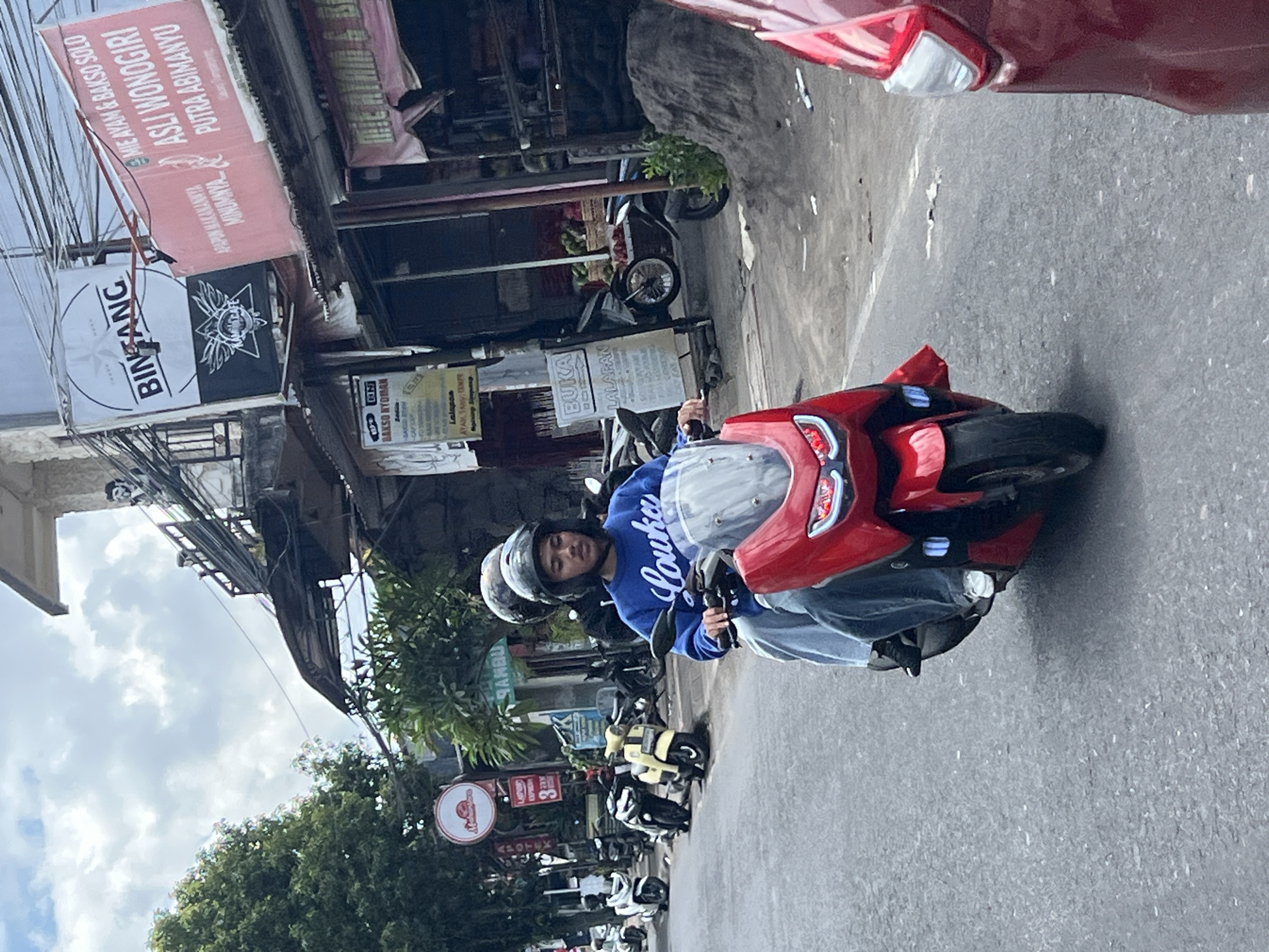 In Payangan, Indonesia, two individuals wearing helmets are riding on a bright red scooter along a paved road. The driver, a young male wearing a blue long-sleeved shirt and light blue jeans, is in the foreground. He is looking forward, seemingly focused on the road ahead. Behind him, another person, partially obscured, is also seated on the scooter. The scooter has a prominent windshield and illuminated headlights.
The road is lined with various establishments, including shops with signage indicating food and drinks. One sign, in red, prominently displays "MIE AYAM & BAKSO SOLO ASLI WONOGIRI PUTRA ABIMANYU." Another white sign reads "BINTANG." Several other motorcycles and scooters are parked along the side of the road. Lush green trees and vegetation are visible in the background, suggesting a tropical environment. The sky above is cloudy, with patches of blue peeking through. The overall scene depicts everyday street life in a bustling area of Payangan.