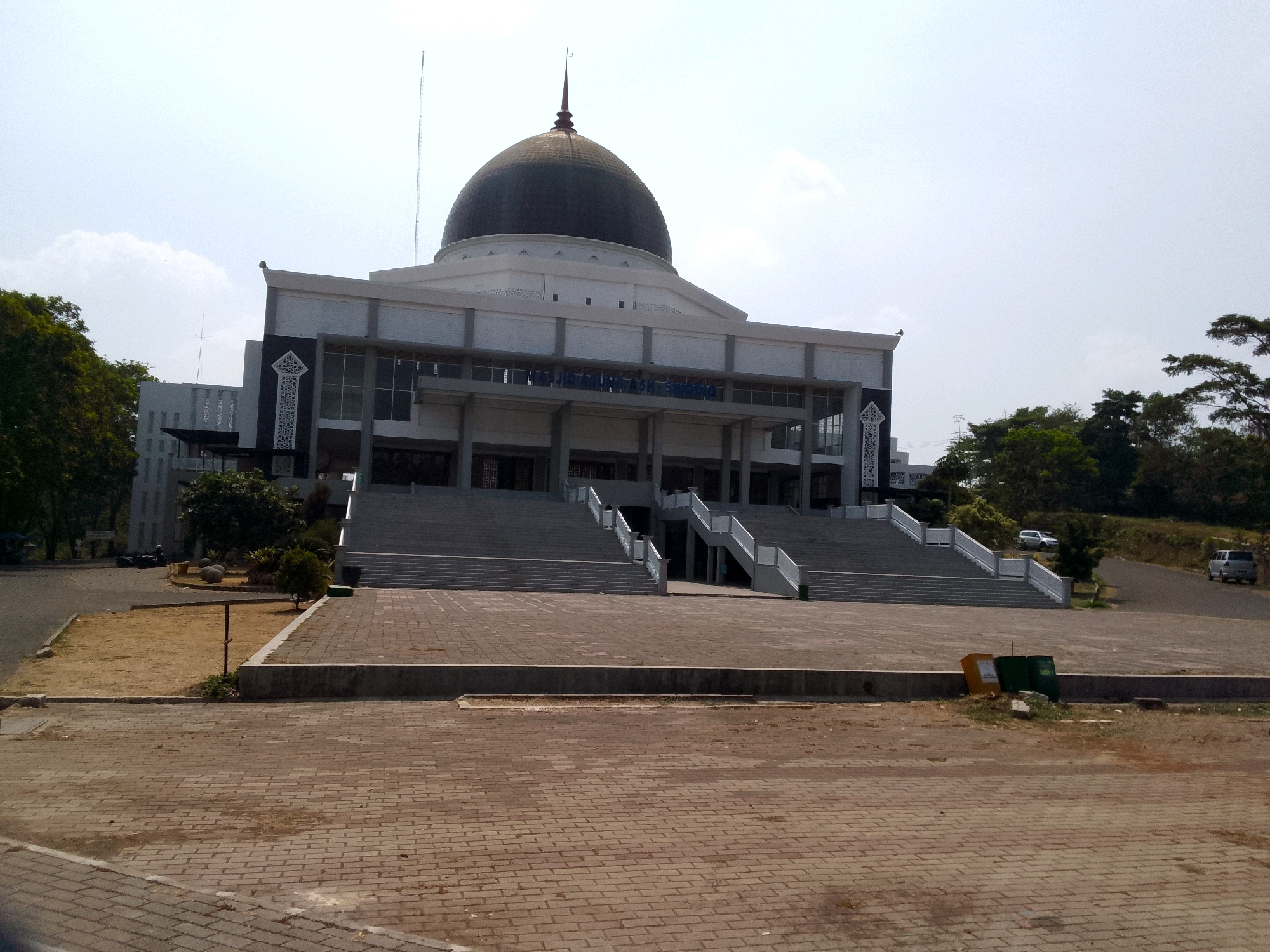 The image depicts the exterior of a large, modern building with a prominent dome, situated in Pasirhalang, Indonesia. The building appears to be a public institution, possibly a government office or a university. A wide, paved plaza with brickwork dominates the foreground, leading up to a grand staircase that ascends to the building's entrance. The architecture features clean lines, grey concrete columns, and large glass windows. Decorative Islamic geometric patterns adorn the facade.
The surrounding environment is characterized by greenery, with trees flanking the building and lining the pathways. A few vehicles are visible parked in the distance, suggesting the presence of visitors or employees. The sky is clear and bright, indicating a sunny day. The overall impression is one of a well-maintained and imposing structure within a spacious urban setting. There are no people prominently featured in the image, emphasizing the architectural focus.