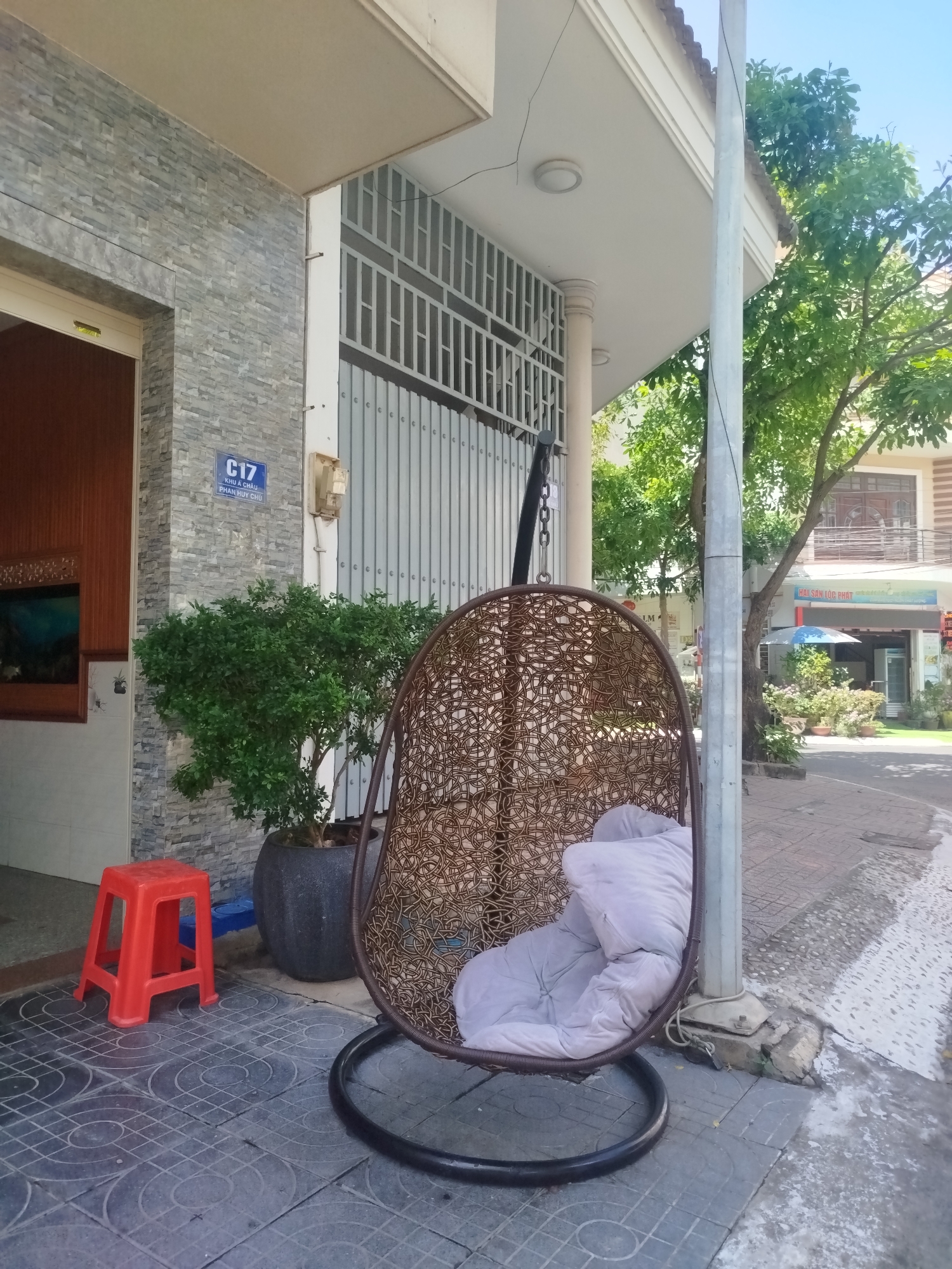 The image depicts a residential street scene in Ho Chi Minh City, Vietnam. The foreground features a hanging chair made of woven material with a light gray cushion, positioned on a tiled sidewalk next to a gray support pole. Behind the chair, a gray tiled building with a 'C17' sign and a plant in a dark gray pot are visible. A small red plastic stool sits nearby. The building has a metal gate and a window with a view into an interior space. Beyond the building and the pole, a street with pedestrians and vegetation leads to other buildings, including one with a 'Hai San Toc Phat' sign. A tree partially obscures the background buildings. The day appears bright and sunny.