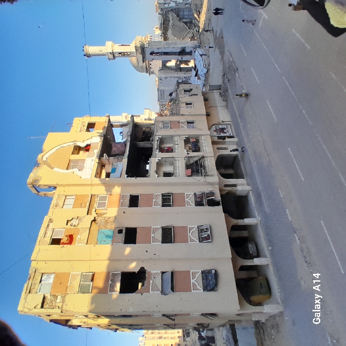 A damaged building stands beside a road, its facade riddled with holes and debris. The structure's upper levels bear the brunt of the destruction, with exposed interiors. Nearby, a mosque shows similar signs of damage, its minaret and dome marked with impact craters. People walk along the street, seemingly unfazed by the surrounding destruction. The scene conveys a sense of resilience amidst devastation, under a clear blue sky.