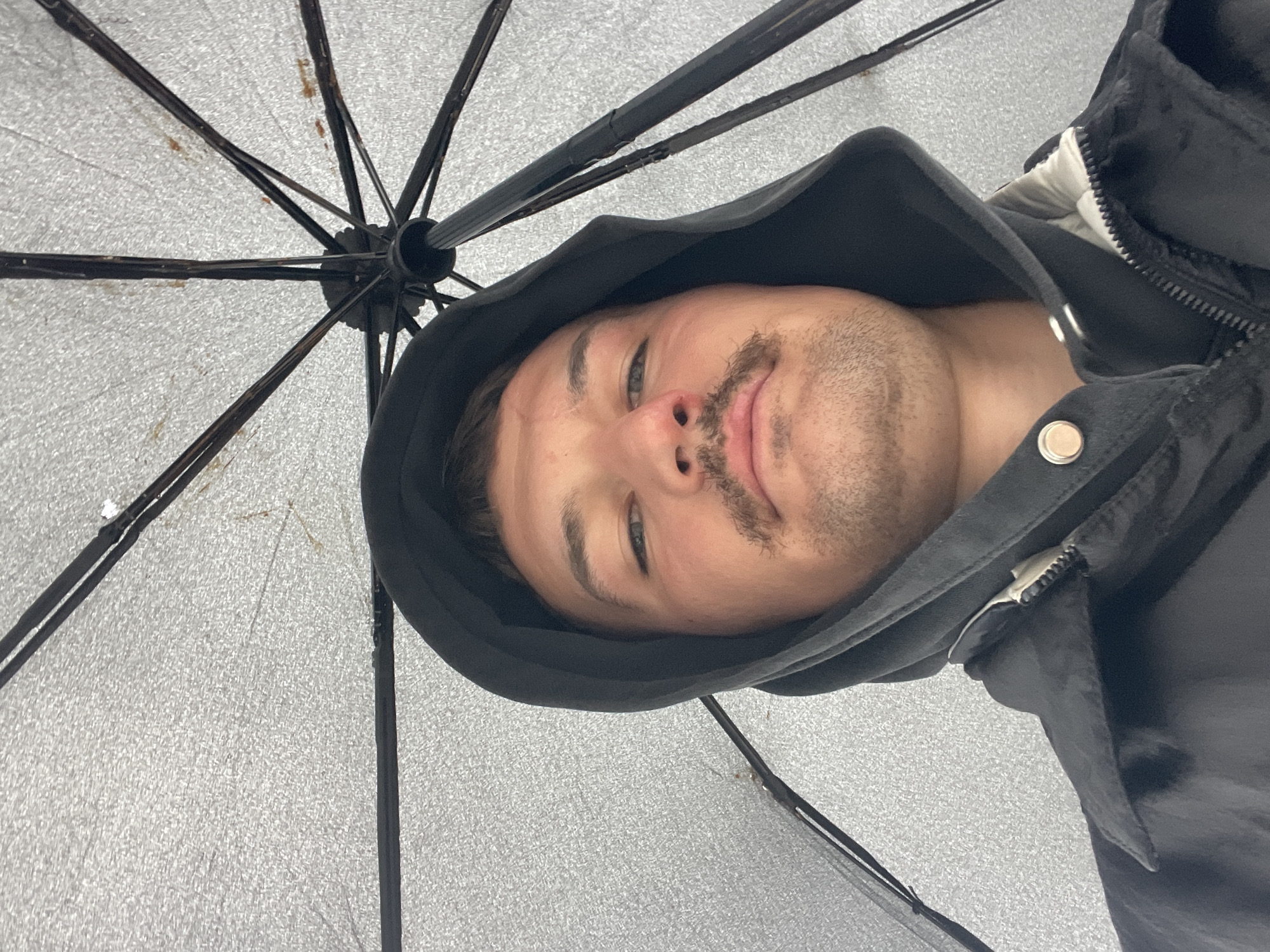 A man with a mustache and wearing a black hooded jacket is holding a silver umbrella, likely outdoors. No specific details indicate the location is Sochi, Russia.