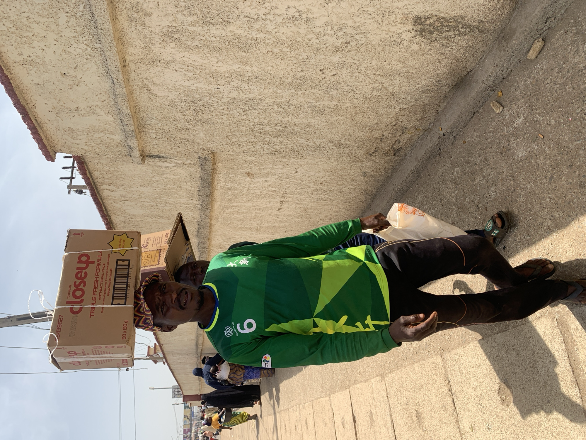 A man wearing a green and yellow soccer jersey with the number "6" is standing on a sidewalk next to a building. He has boxes of "closeup" toothpaste balanced on his head. There are other people in the background. The location appears to be in Jos, Nigeria.