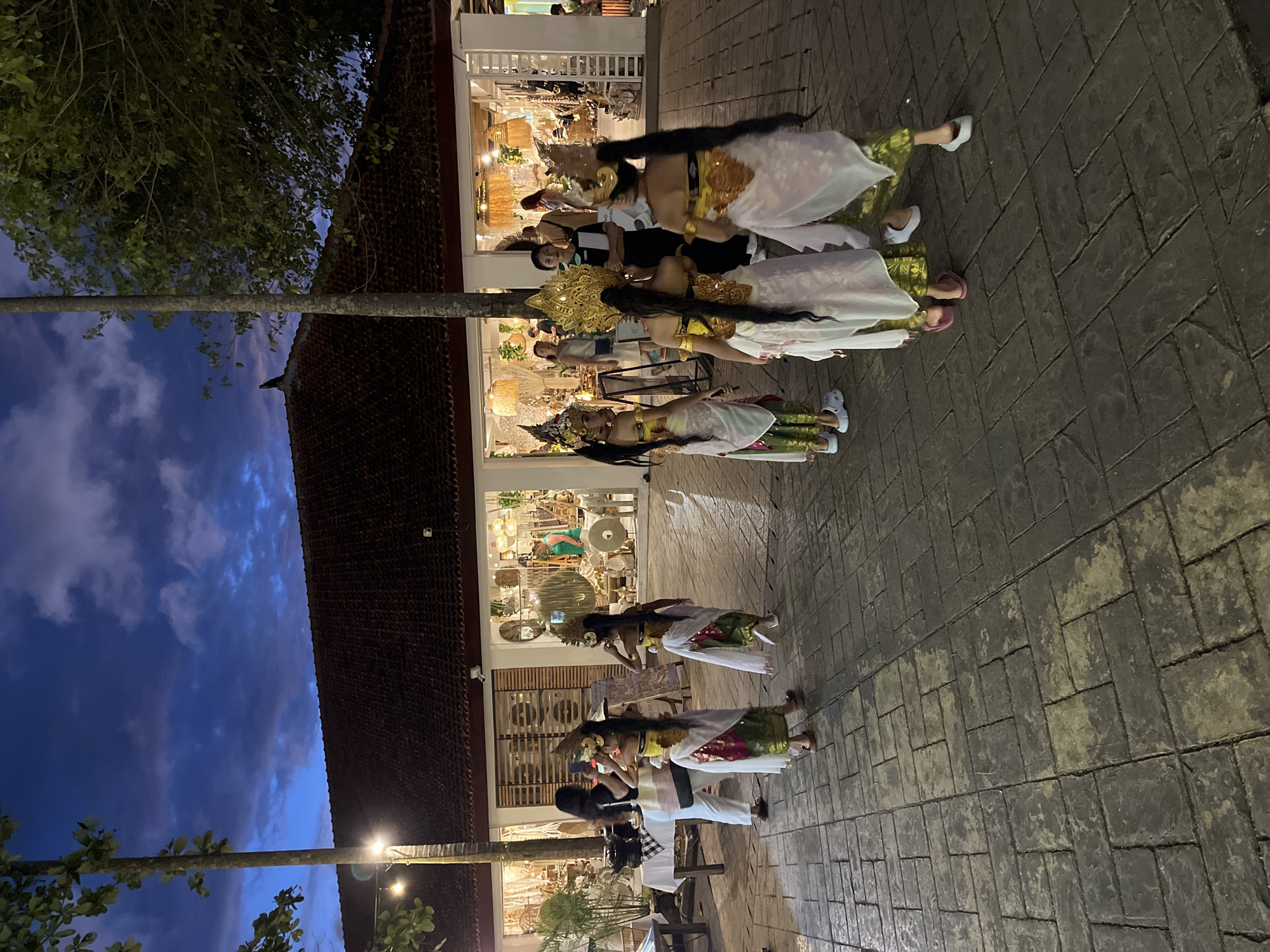 At dusk in Kuta Selatan, Indonesia, a procession of Balinese dancers in traditional attire are situated on a brick-paved street. The dancers are predominantly dressed in white and gold costumes, some wearing elaborate headdresses. They are walking along a row of shops with illuminated window displays. Above, the sky is a gradient of dark blue and hints of twilight. A slender tree trunk is visible on the left side of the frame.