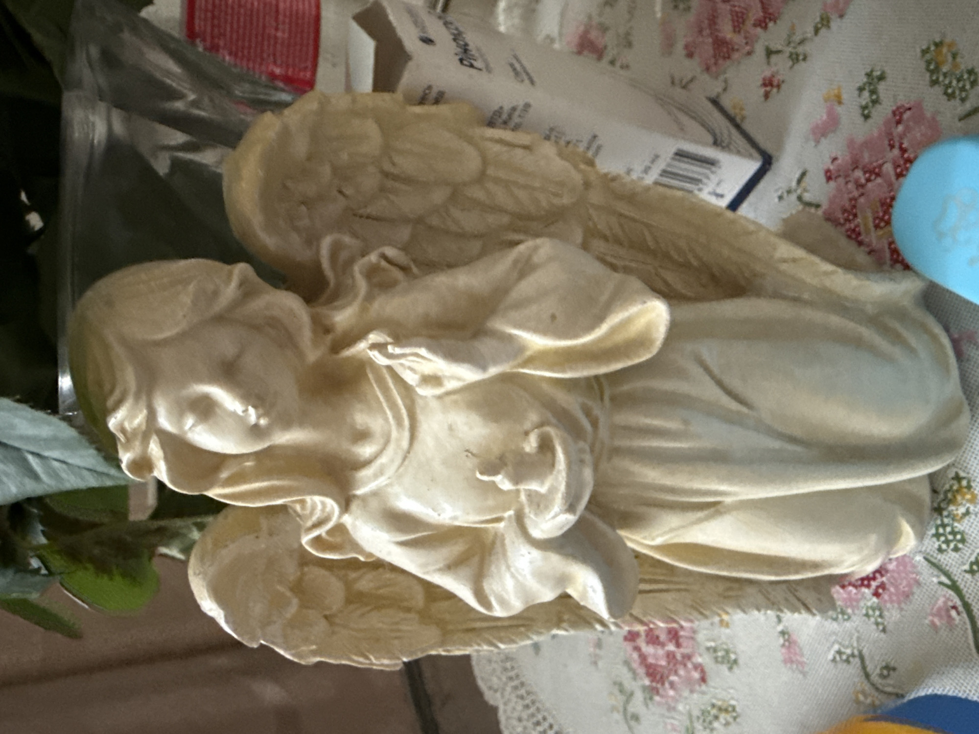 An antique beige angel statue with detailed wings and flowing robes kneels on a floral tablecloth. The statue holds a small, unlit candle in its left hand. The background features green foliage from a plant and a white cardboard box with red accents and text. The lighting is soft, suggesting an indoor environment.