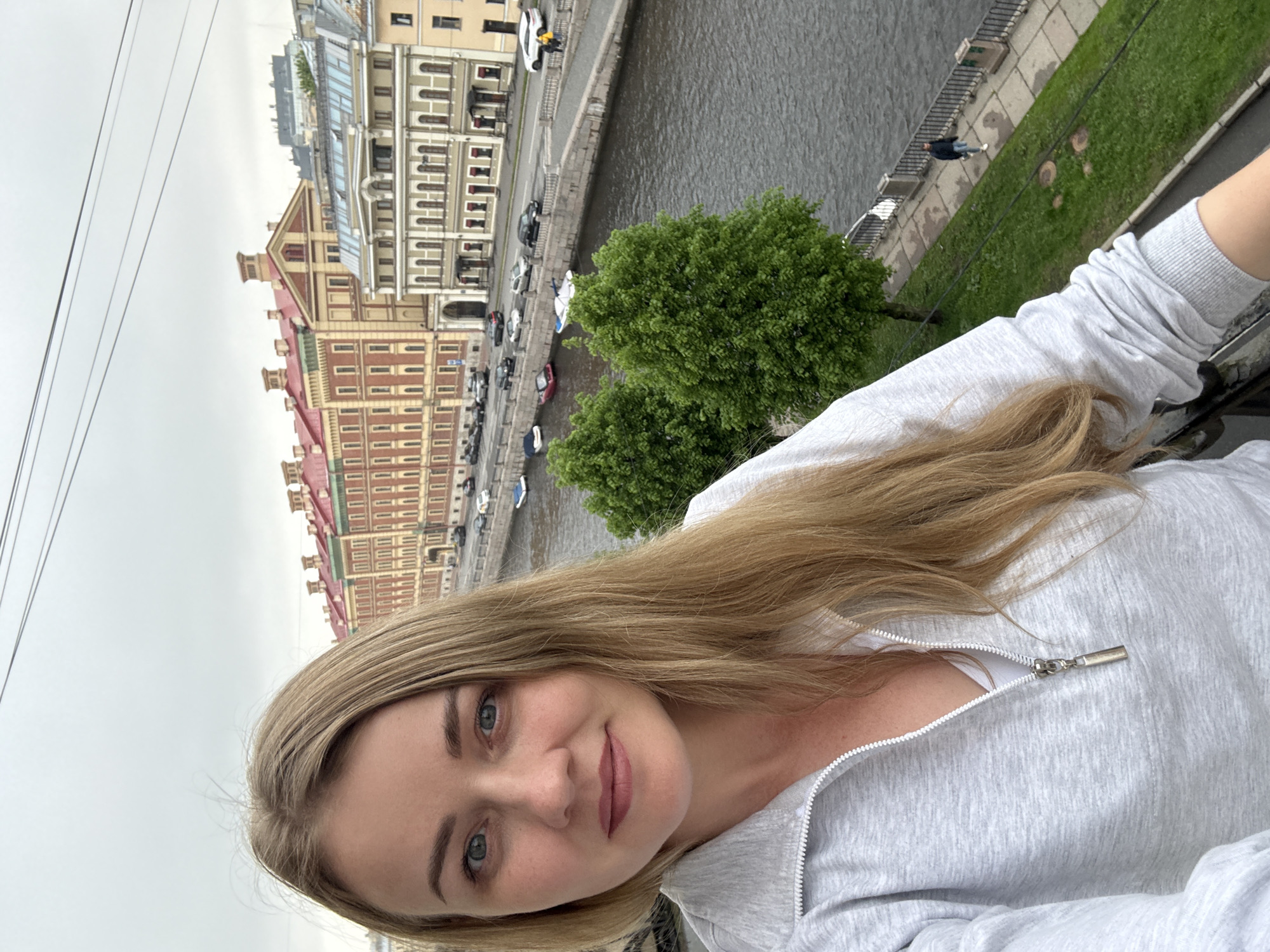 A woman with blonde hair wearing a light grey zipped jacket is taking a selfie. The background shows a view of Saint Petersburg, Russia, specifically along a canal or river. Buildings line the water, some with a distinctive red roof. There are cars parked along the canal, and green trees are visible. The sky is overcast. A person can be seen walking along the embankment.