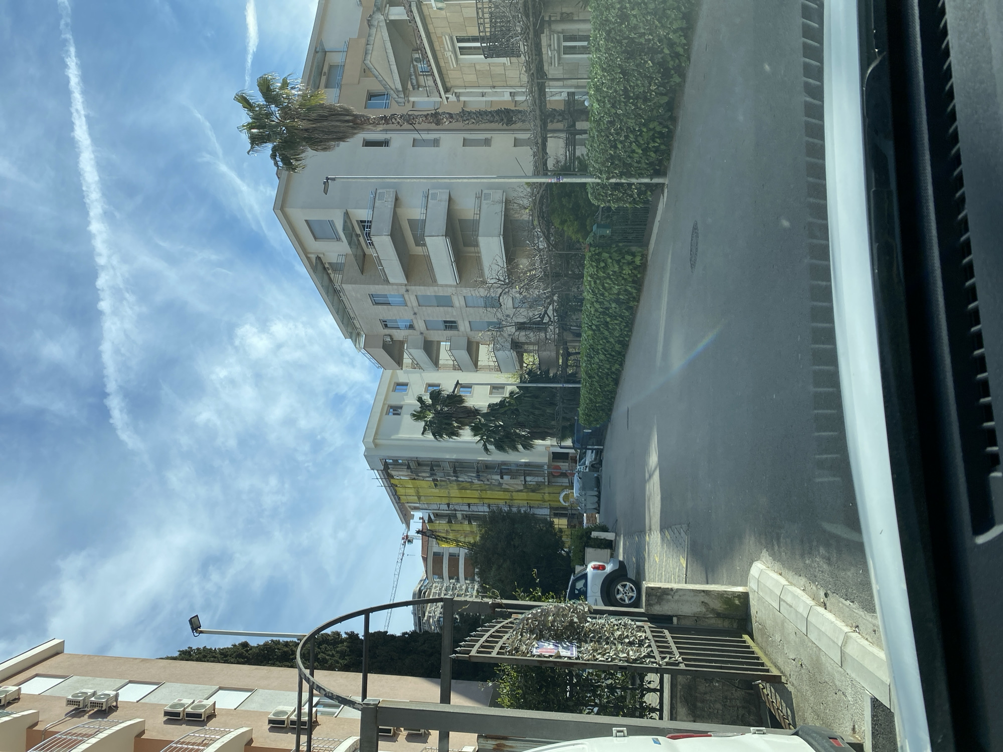 The image depicts a street scene in Budva, Montenegro, taken from inside a vehicle. The view is oriented towards residential buildings lining the street. The buildings are multi-story with balconies, typical of urban architecture in the area. One building has scaffolding present, indicating ongoing construction or renovation. Palm trees are visible interspersed with the buildings. On the street, parked cars are present, along with greenery and streetlights. The sky is blue with contrails. There are metal fences in the foreground obscuring the lower part of the view.