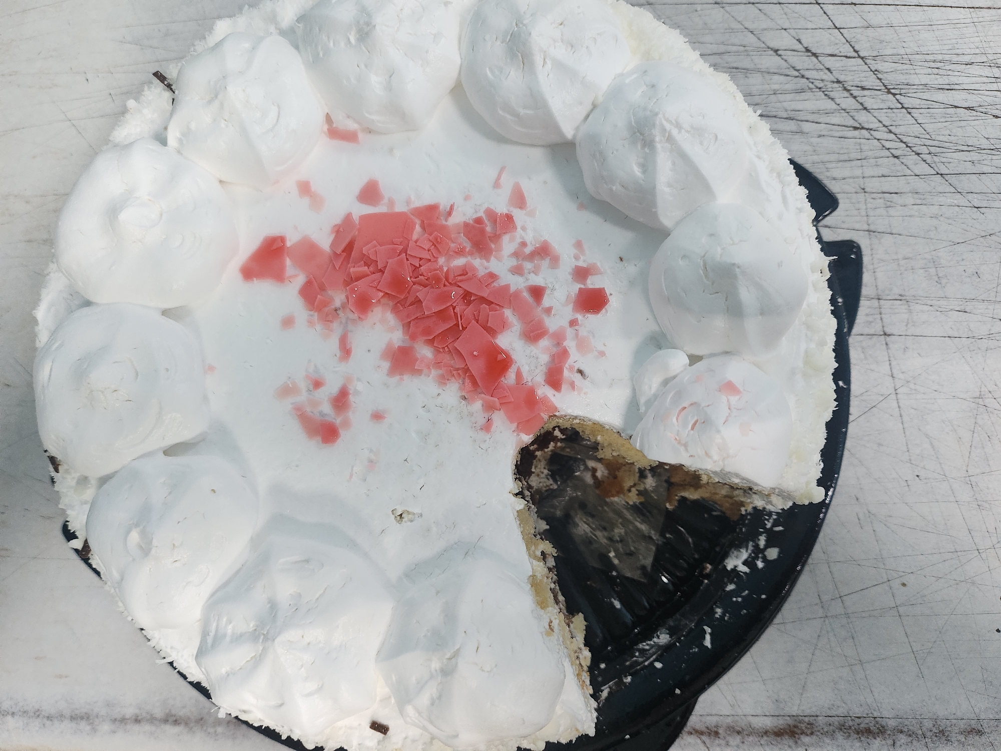 This overhead shot features a round cake, partially eaten, sitting on a textured surface. The cake is predominantly covered in a thick, white frosting or whipped cream, piped into large, soft dollops around its perimeter and smoothed across the top. In the center of the white topping, numerous irregular, shard-like pieces of pink or light red candy are scattered as decoration.
A triangular slice has been cut from the cake, revealing its interior. The exposed layers show a strikingly dark, almost black, and somewhat glossy filling or cake core, which appears to be a rich chocolate or dark mousse. Beneath this dark layer, a lighter, crumbly brown base, possibly biscuit or sponge, is visible. The cake rests on a round, black plastic or cardboard cake board.
The cake is placed on a light-colored, likely white or light grey, surface that serves as the background. This surface is heavily marked with numerous scratches, lines, and scuff marks, suggesting it is a well-used countertop or table, possibly in a kitchen or serving area. No people are visible, and no text is discernible on the cake or in the background. The scene depicts a cake that has recently been served and enjoyed. The location context is Krasnogorskoye, Russia, though no specific visual cues in the image directly indicate this.
