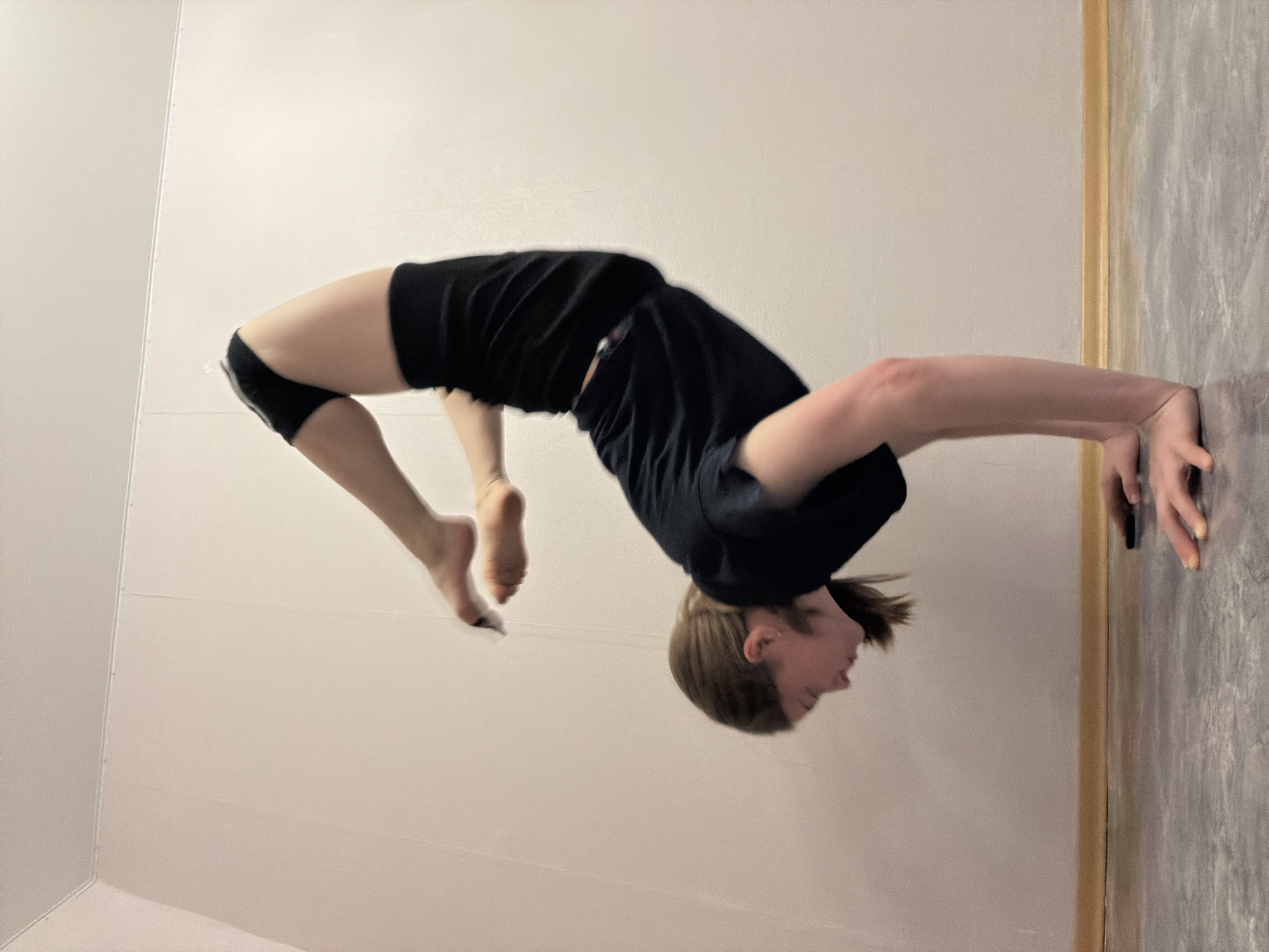 A person is captured in a backbend pose in what appears to be an indoor space. The person is wearing dark shorts and a dark t-shirt, with their hands pressed against a wall. The background consists of a light-colored wall and a darker wall with a trim separating the two. The person's feet are off the ground. The location, Perm, Russia, cannot be verified based on the image content alone.