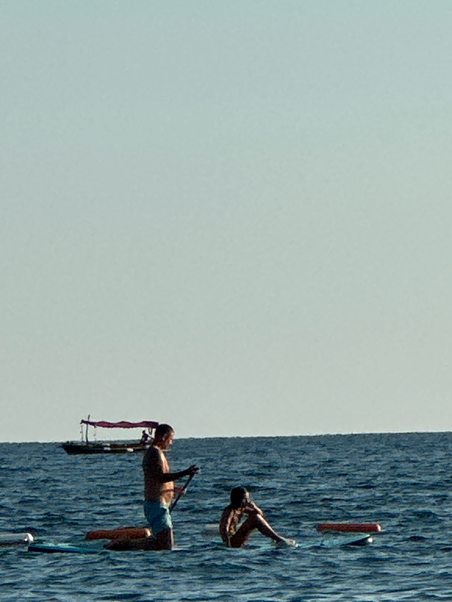 In the waters off Boreti, Montenegro, two individuals are engaged in paddleboarding. A man, standing on his paddleboard, holds a paddle. Another person, seated on their paddleboard, faces away from the viewer. In the background, a boat with a canopy is visible. The sea is calm, and the sky is clear and pale.