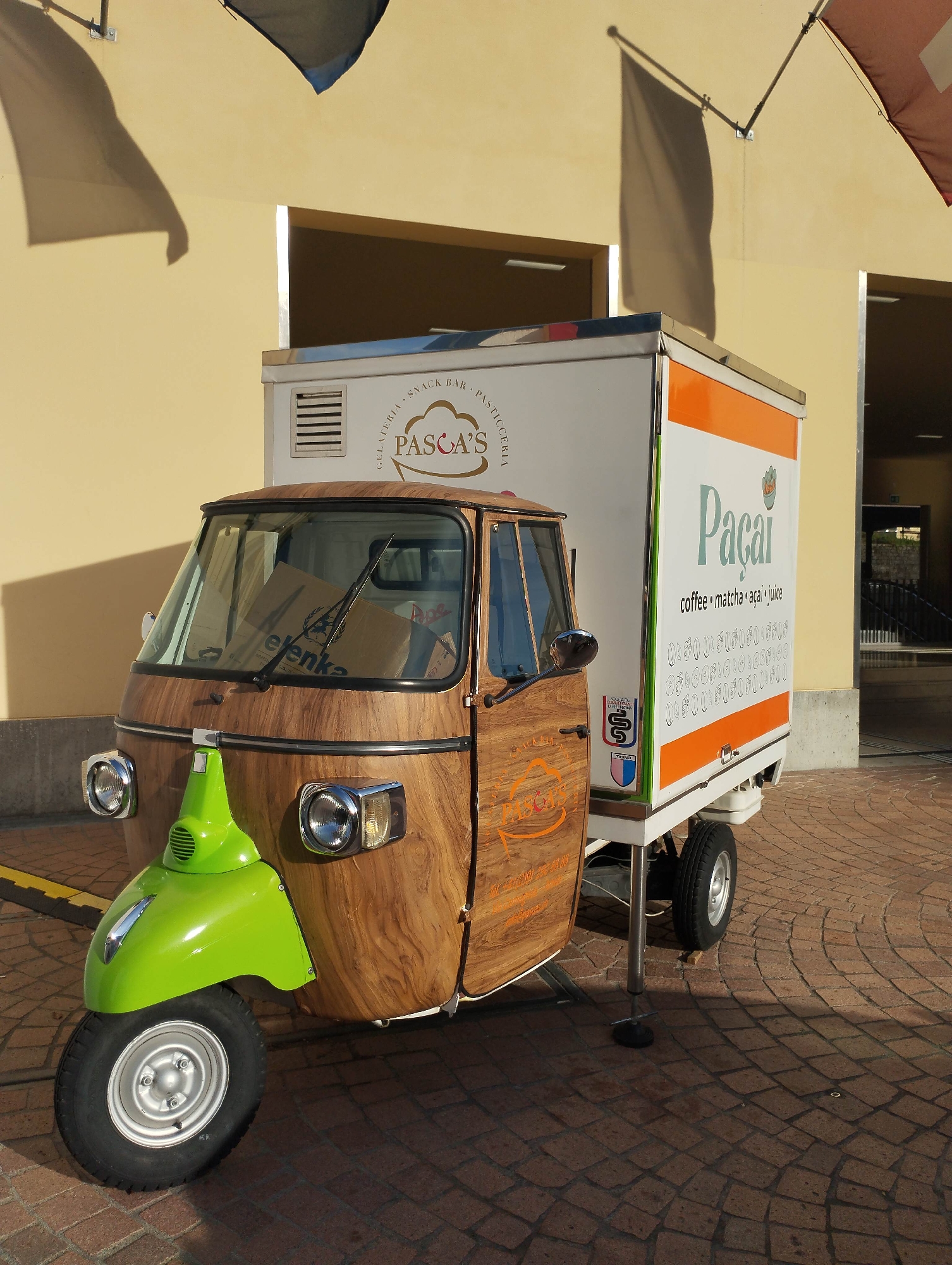 A unique three-wheeled vehicle, resembling a Piaggio Ape, is parked outdoors, functioning as a mobile food and beverage stand. The vehicle's cab features a distinctive wood-grain finish, with a vibrant lime green front fender. Chrome headlights and turn signals are visible on the front of the cab. Inside the windshield, several cardboard boxes can be seen, one bearing the text "elenka."
Attached to the rear of the cab is a white cargo box, serving as the main vending area. The front panel of this box displays "GELATERIA • SNACK BAR • PASTICCERIA" arranged in an arc above a cloud-shaped logo with "PASCA'S" and a red accent. The side of the cargo box prominently features "Paçaí" in large green letters, with "coffee • matcha • açai • juice" written below it, followed by a pattern of small, circular graphics. The driver's side door of the cab also has a "PASCA'S" logo with additional, smaller text. A small crest or emblem is visible on the side of the cargo box, near the cab door. Two extendable support legs with black bases are deployed from the cargo box, stabilizing the vehicle.
The scene is set on a patterned reddish-brown cobblestone ground. In the background stands a light yellow building with recessed openings. Long, distinct shadows, cast by unseen objects (possibly flags or awnings) on the building and by the vehicle itself, indicate bright, direct sunlight, suggesting it is daytime, possibly morning or late afternoon. Flags or banners are also visible at the top of the frame, attached to the building. No people are present, but the vehicle is clearly set up for commercial operation.