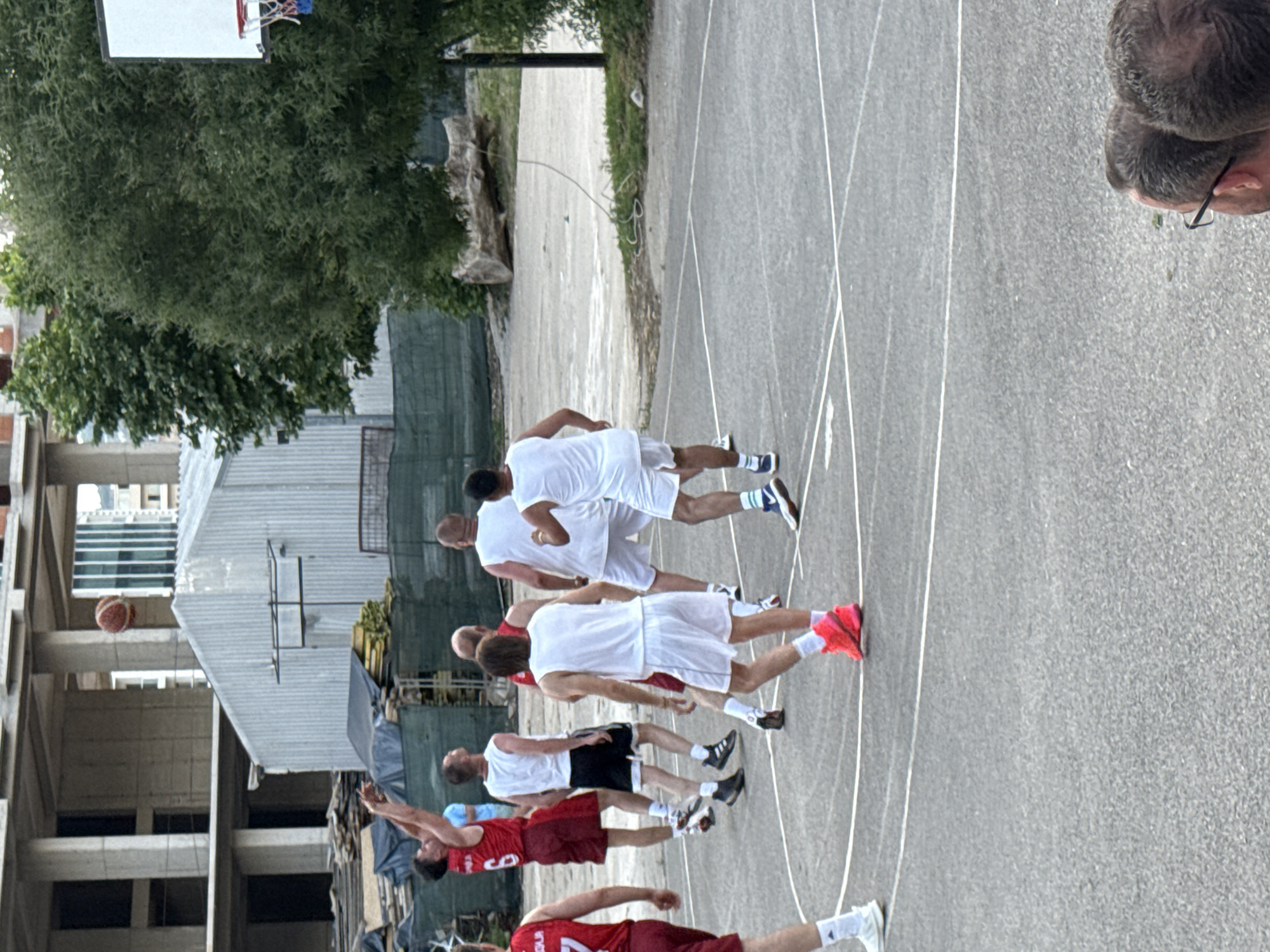 A group of men are playing basketball on an outdoor court. One man in a red jersey with the number 6 is in the air, shooting a basketball. Several other men, mostly in white jerseys and shorts, are positioned on the court. The court is paved and has white lines marking its boundaries. In the background, there is a large, unfinished concrete building, a corrugated metal structure, trees, and a basketball hoop. The overall scene suggests a casual game of basketball taking place in an urban or semi-urban environment, possibly in Budva, Montenegro, given the context of construction and outdoor courts commonly found in the region.