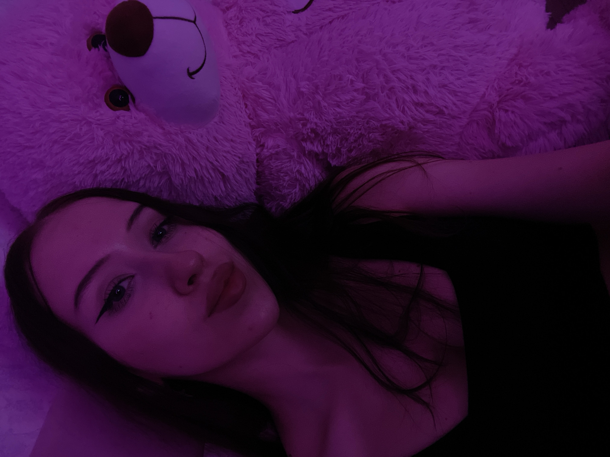 A young woman with dark hair and winged eyeliner poses for a selfie next to a large, plush pink teddy bear. The scene is illuminated by purple lighting, casting a strong color cast on both the woman and the bear. The woman is wearing a black tank top. The teddy bear has large brown eyes and a stitched smile. The image appears to have been taken indoors, possibly in a bedroom. There are no discernible landmarks or environmental cues to suggest a specific location within Votkinsk, Russia.