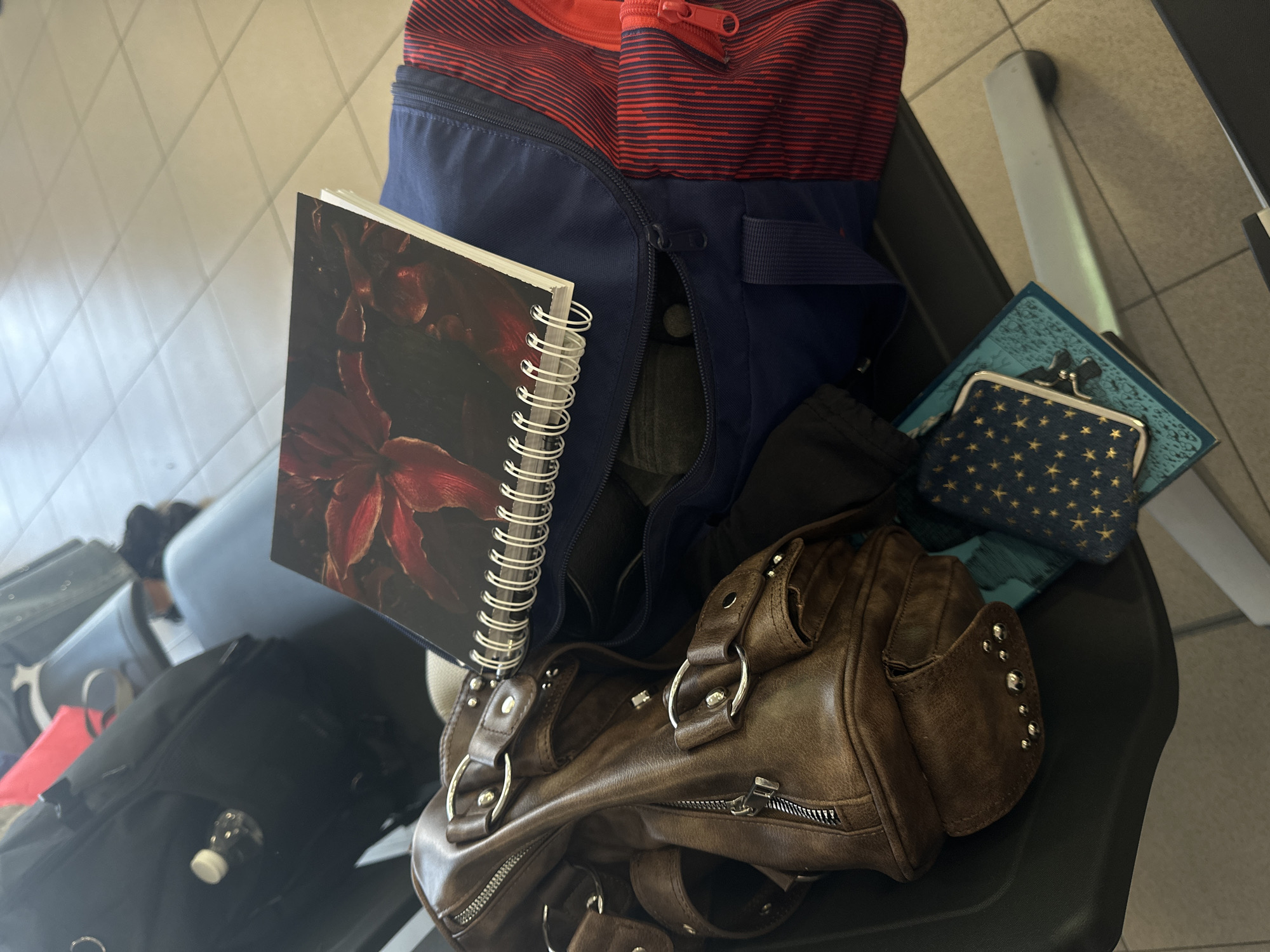 This is a close-up, high-angle shot of several bags and a notebook, likely staged or packed for travel. The objects are resting on a dark, possibly plastic chair. In the foreground, a brown leather handbag with silver hardware is prominent, showing zippers and decorative studs. To the left of the handbag, a blue and red striped duffel bag is partially open, revealing some dark fabric inside. A spiral-bound notebook with a dark floral cover is propped against the duffel bag. Further back, a small blue coin purse adorned with gold stars is visible, resting on top of another blue and green striped item. In the background, other dark-colored bags and what appears to be a small plastic bottle with a white cap can be seen. The scene is indoors, with a tiled floor visible in the background. The lighting suggests it could be daytime, but there are no specific clues about the time of day or weather. There is no visible text in the image. The arrangement of the items suggests preparation for a journey or a moment of rest during travel, possibly at an airport or train station, given the utilitarian style of the seating.