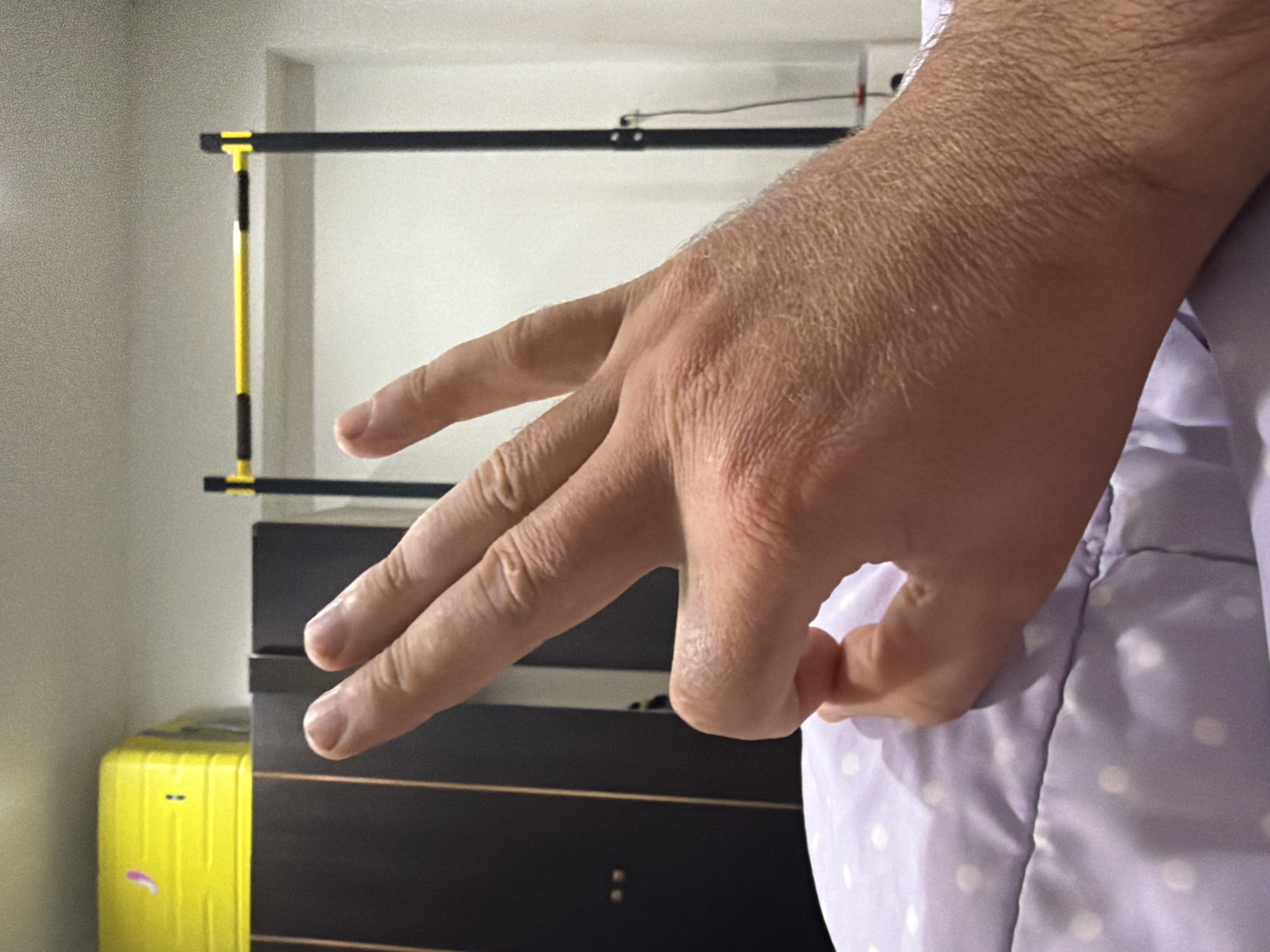A close-up, head-on shot shows a person's right hand in the foreground, making an "okay" gesture with the thumb and index finger touching, and the other three fingers extended upwards. The hand is positioned over a purple and white polka-dotted surface, possibly bedding. In the background, on the left, is a dark brown wardrobe with a yellow suitcase placed on top. To the right of the wardrobe, a black and yellow pull-up bar is visible against a white wall. The scene appears to be indoors, in a bedroom or living space. The lighting is soft, casting subtle shadows.