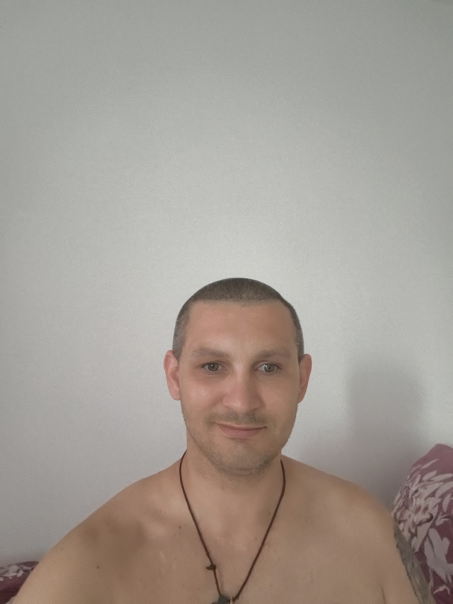 A man with short hair and a necklace is pictured from the shoulders up. He is looking directly at the camera with a slight smile. The background is a plain, light-colored wall. The man is positioned in the center of the frame.